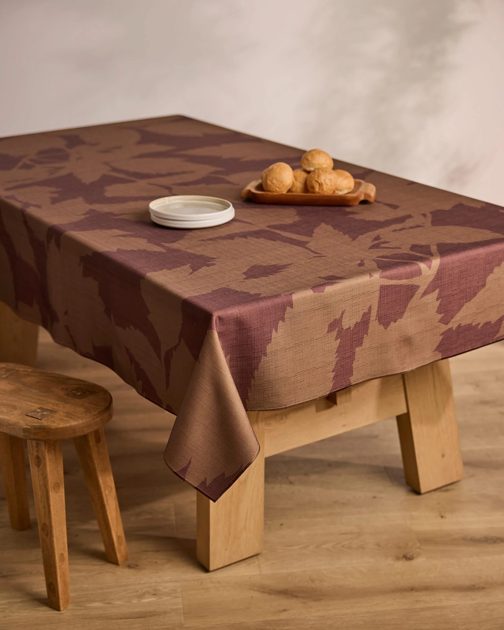 The Geometry Rustle table, draped in a brown and mauve stain-resistant cloth, features a small plate, a tray of bread rolls, and comes with a matching wooden stool—all set on a light wood floor.