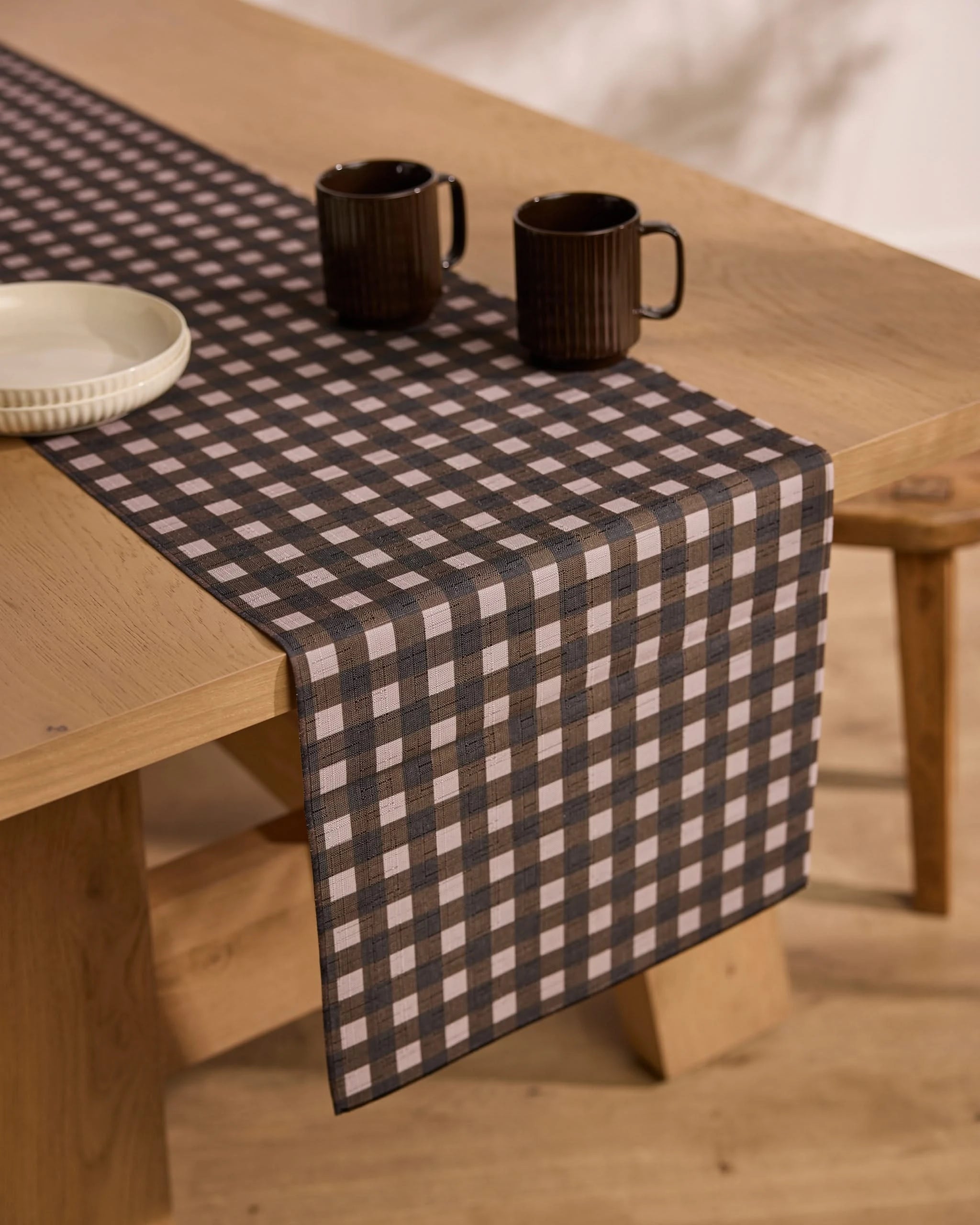 The Deep Hearth table runner by Geometry, in black and white checks, is styled on a wooden dining table with two dark ceramic mugs and a white plate for a cozy, rustic vibe.
