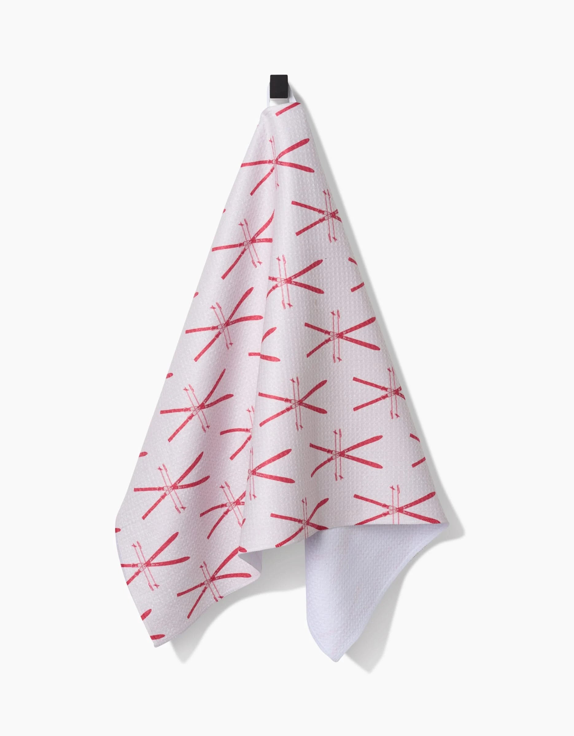 The Becki Owens Vintage Ski tea towel features a red crossed skis pattern on white Geoweave fabric and hangs on a black hook against a white background—an eco-friendly, stylish addition to any kitchen.