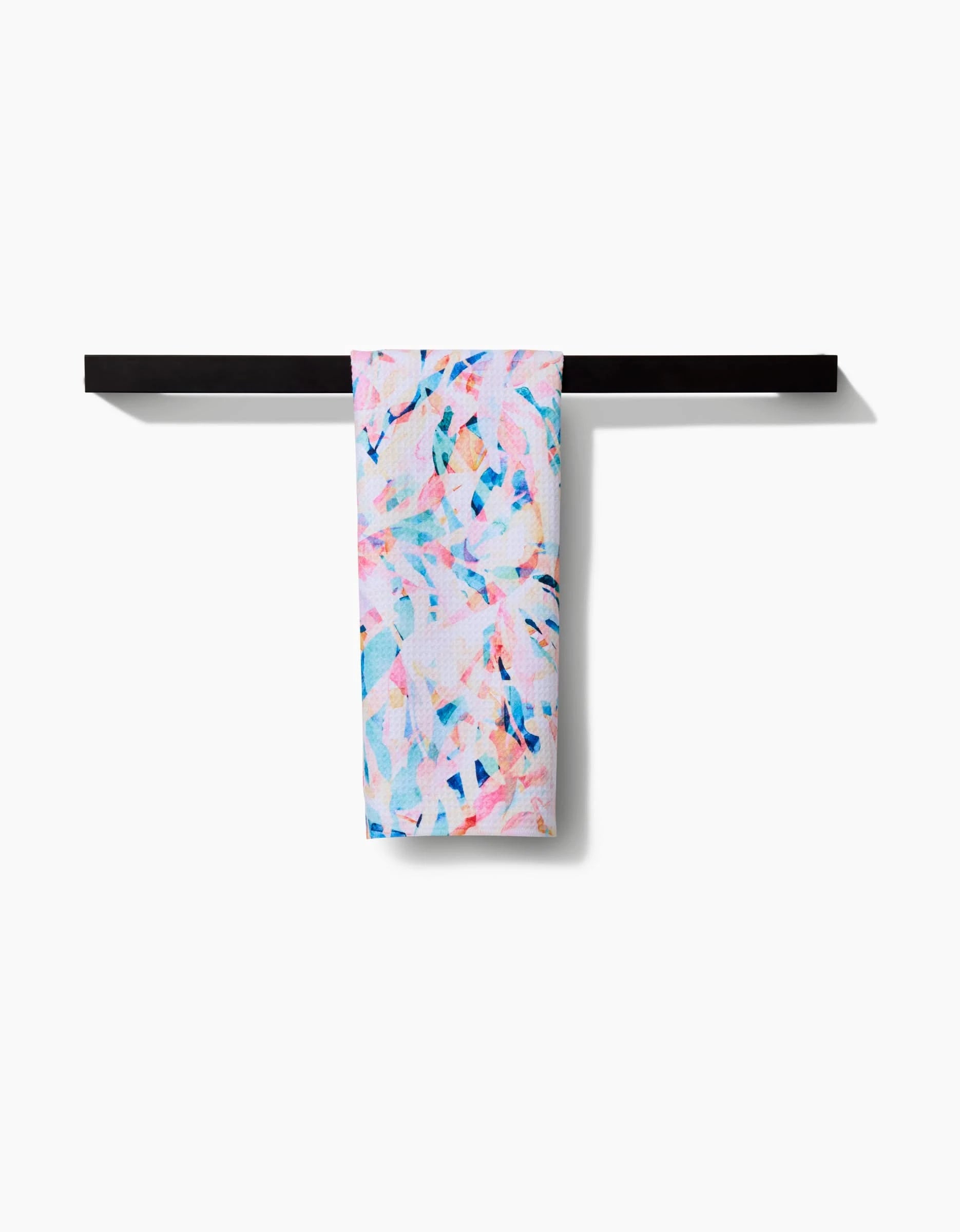 The Rachel Parker Abstract Jungle tea towel, featuring a colorful pastel pink, blue, and white abstract pattern, hangs neatly on a sleek black towel bar against a white background.