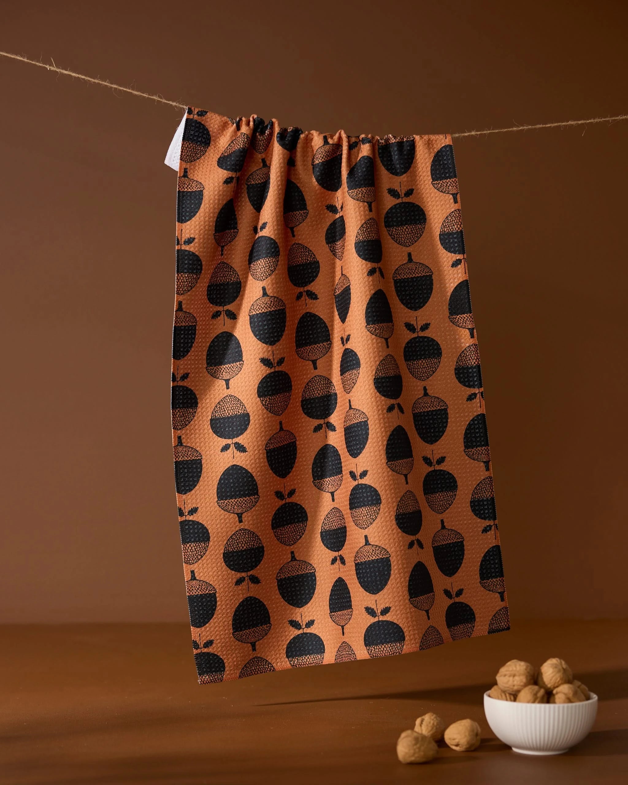 The Acorn Noir kitchen tea towel by Geometry, featuring a black acorn pattern and made from recycled materials, hangs on a string above a small white bowl of walnuts and loose nuts, set against a brown background.
