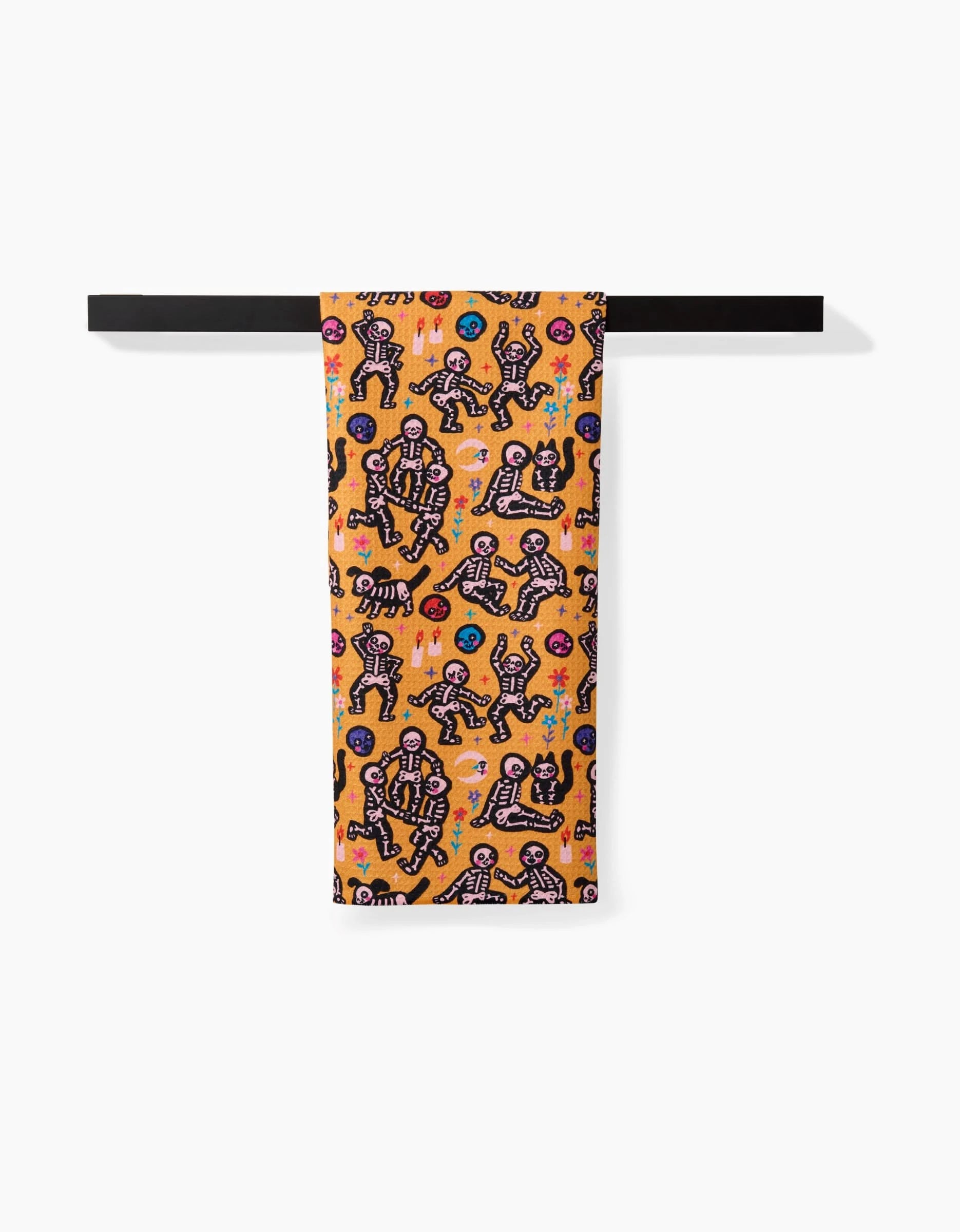 The So Lazo After Party tea towel, featuring playful abstract human and animal figures and geometric shapes in black, blue, pink, and white on a bright orange background, hangs on a horizontal black rod against a white backdrop.
