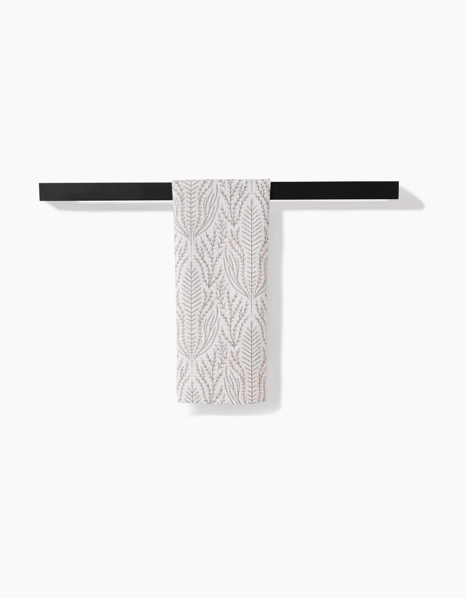 The Amberlin tea towel by Amy MacCready, featuring a light brown leaf pattern on white and made from recycled materials, is neatly folded over a sleek black towel bar against a plain white background.