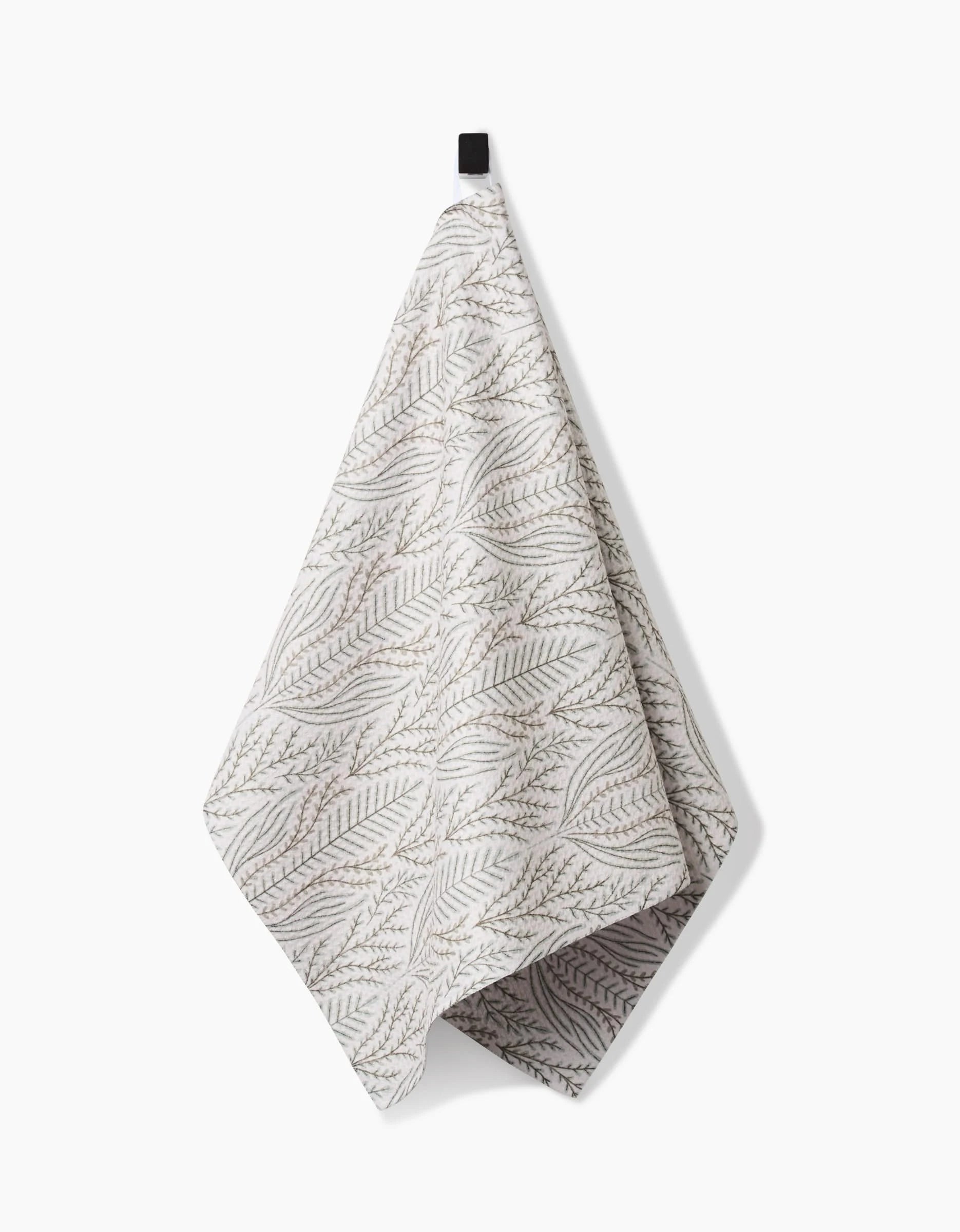 The Amberlin tea towel by Amy MacCready, made from recycled materials, features a subtle botanical leaf pattern in a light color and hangs from a black loop against a white background.