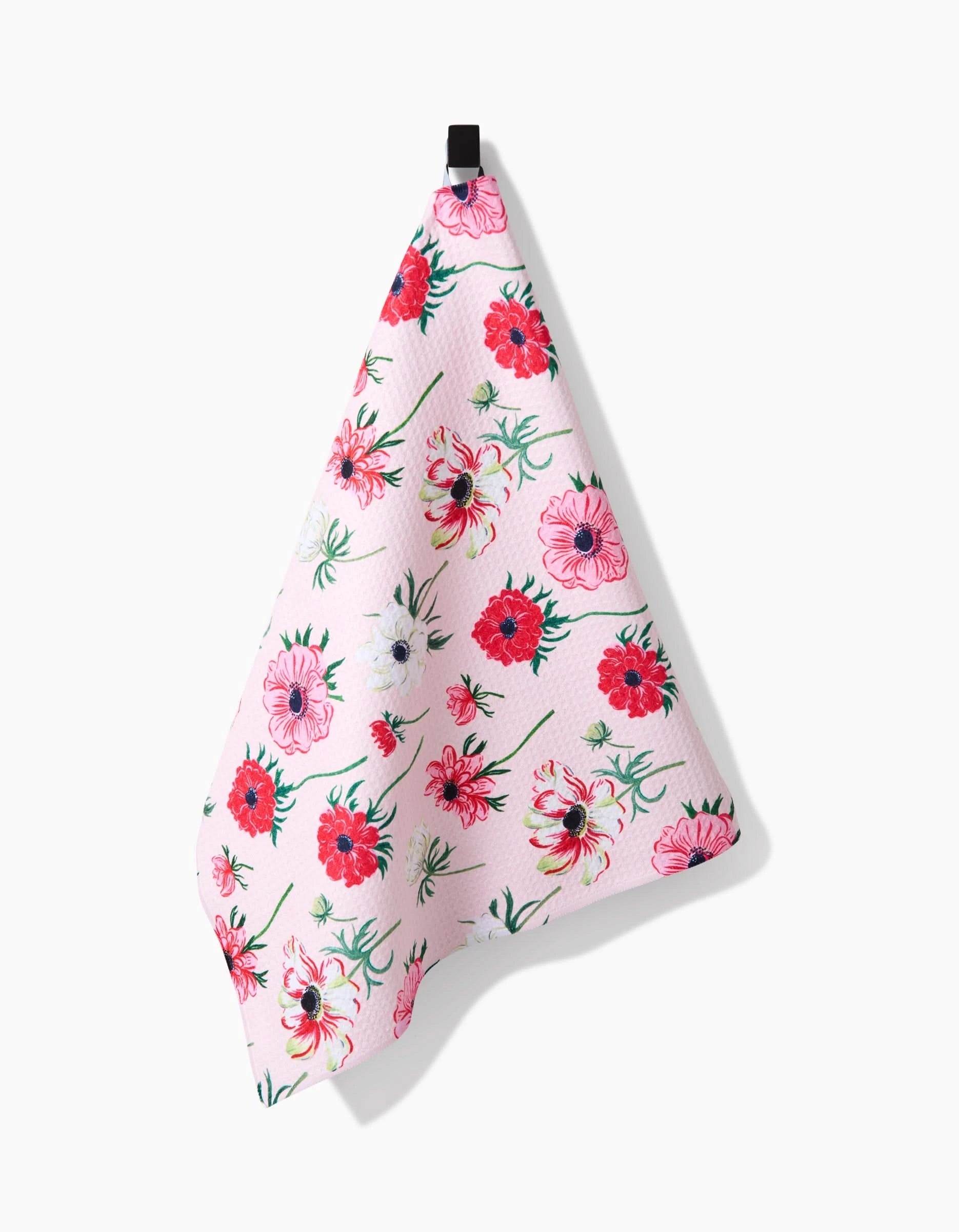 The Linda Fox Anemone Garden Flowers tea towel, in light pink with red, pink, and white floral patterns and made from recycled materials, hangs from a black hook against a plain white background.