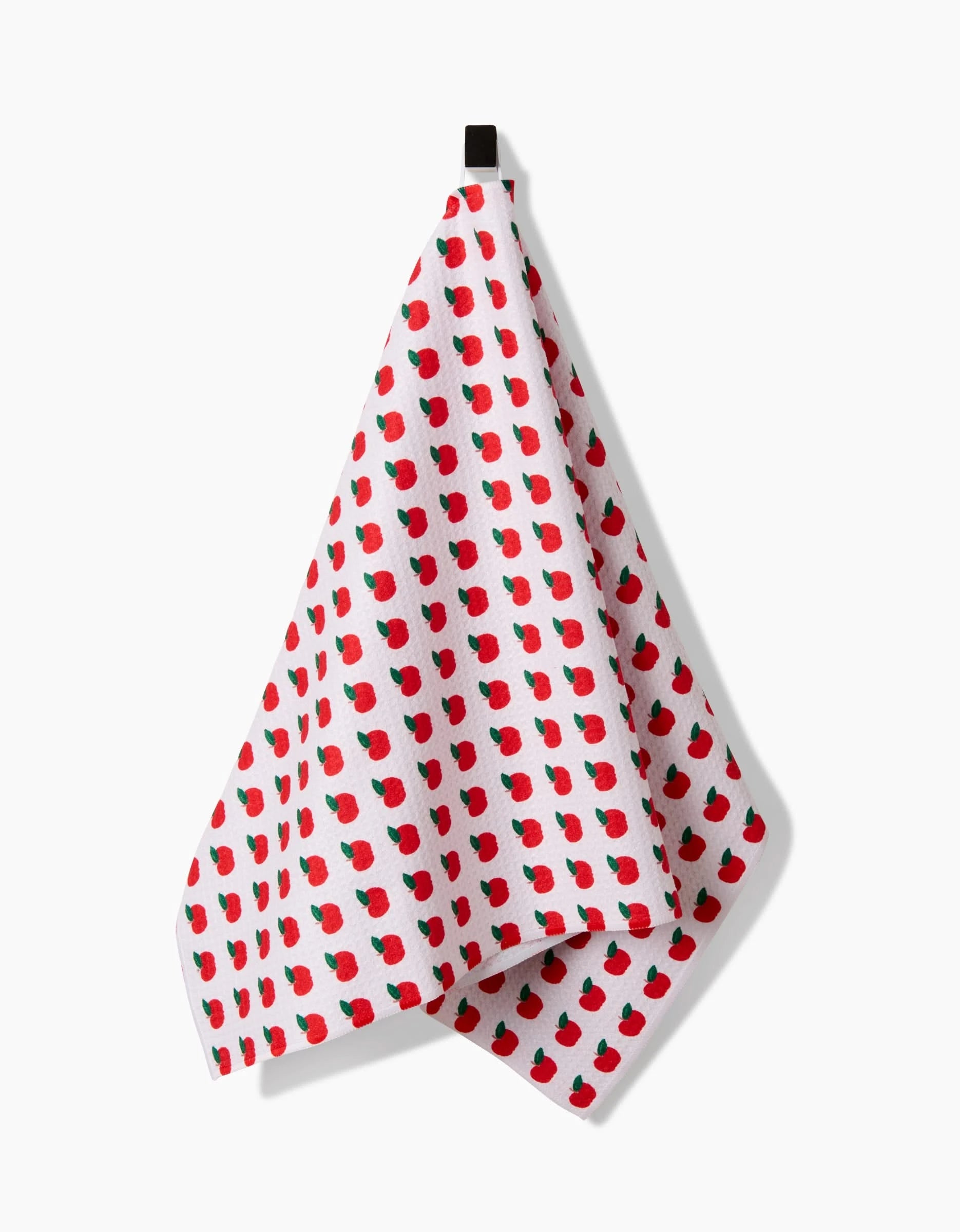 The Apple Grove tea towel by Brittany Jepsen, made from recycled materials, features a repeating pattern of small red apples and green leaves and is shown hanging from a black hook against a plain white background.