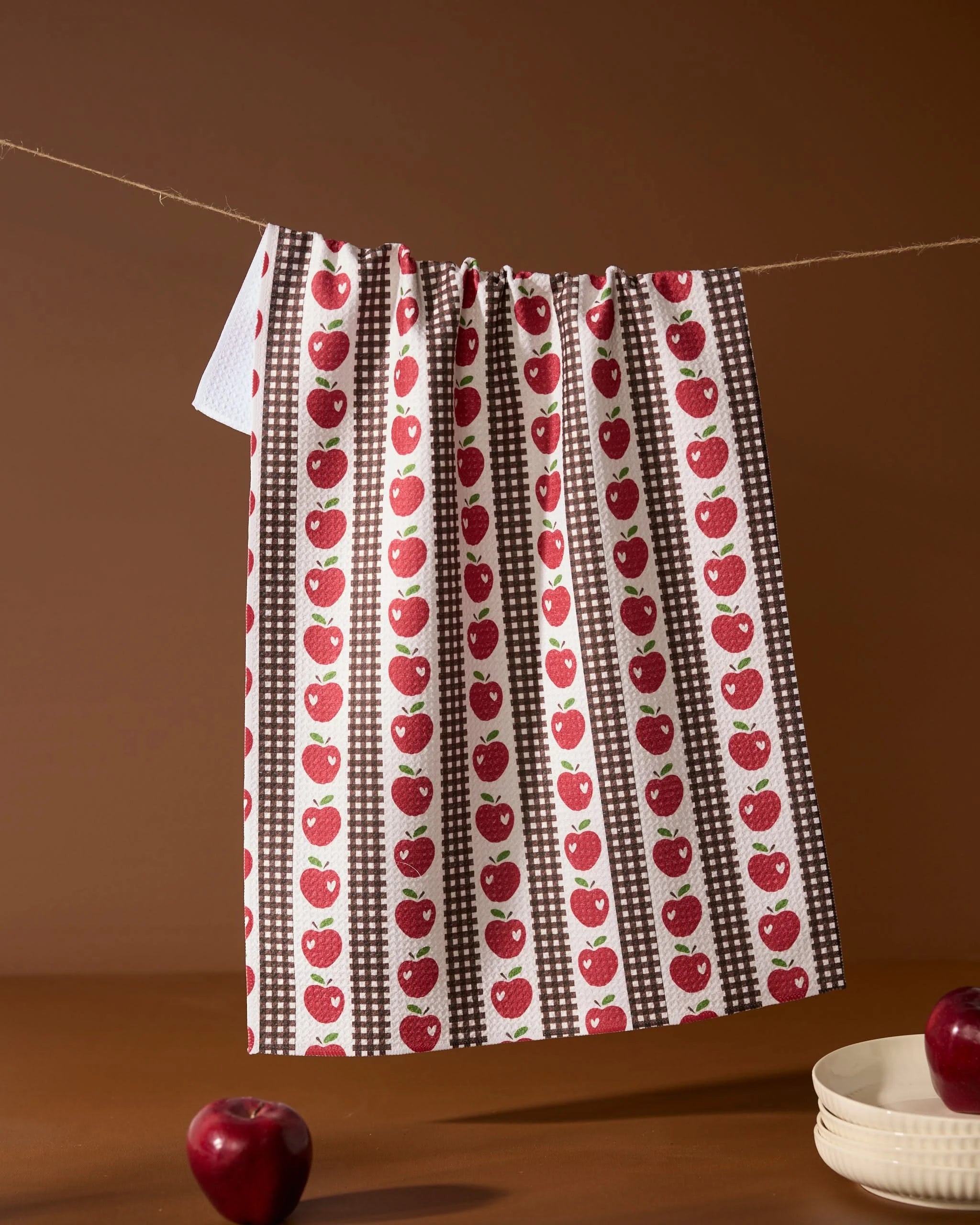 The Geometry Apple Picking Day kitchen towel, featuring red apples and brown checkered stripes, hangs above stacked white plates and apples on a brown surface with a matching background.
