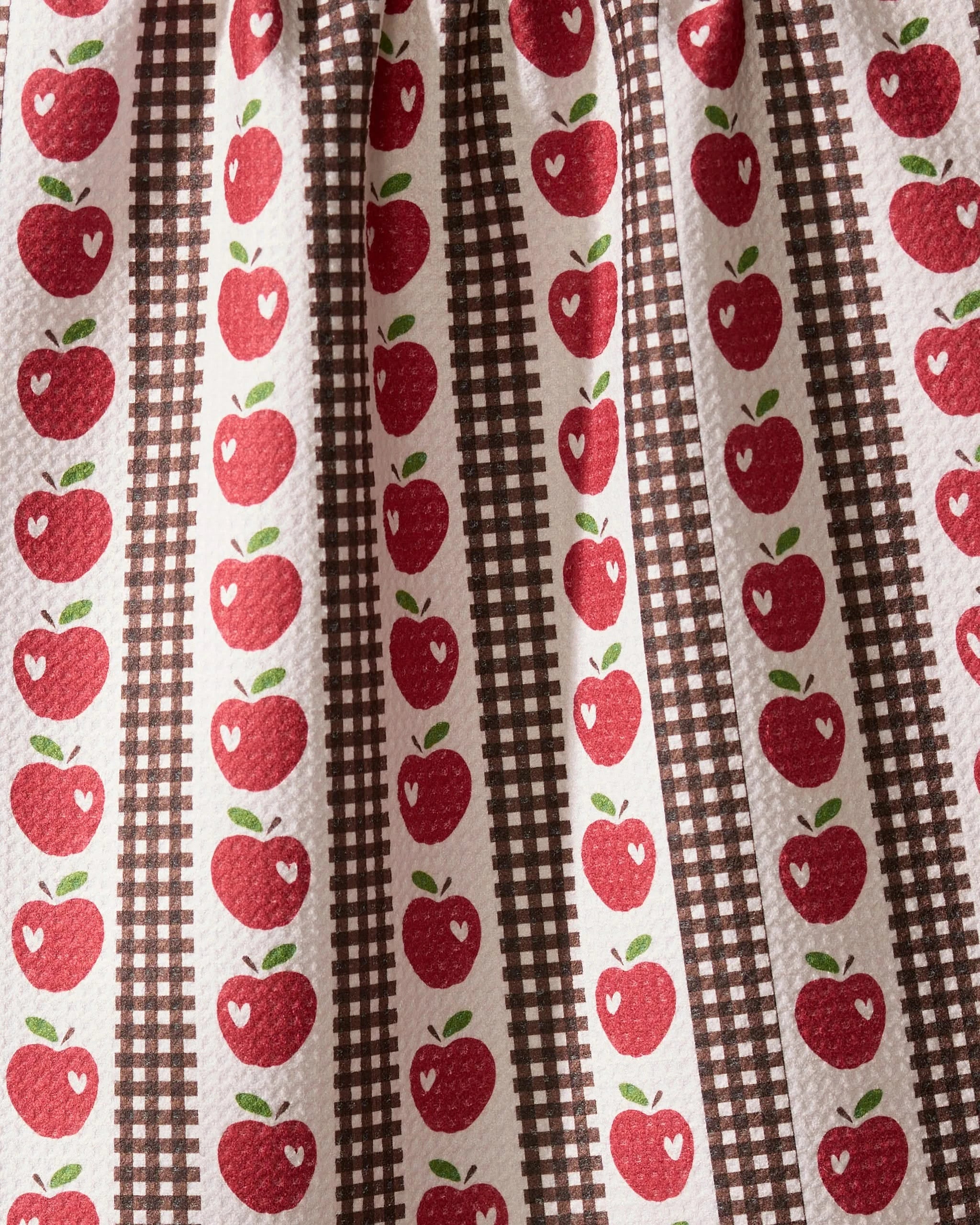 The Geometry “Apple Picking Day” tea towel features red apples with white highlights in vertical rows, alternating with brown plaid stripes on a textured white background for a charming, vintage country kitchen look.