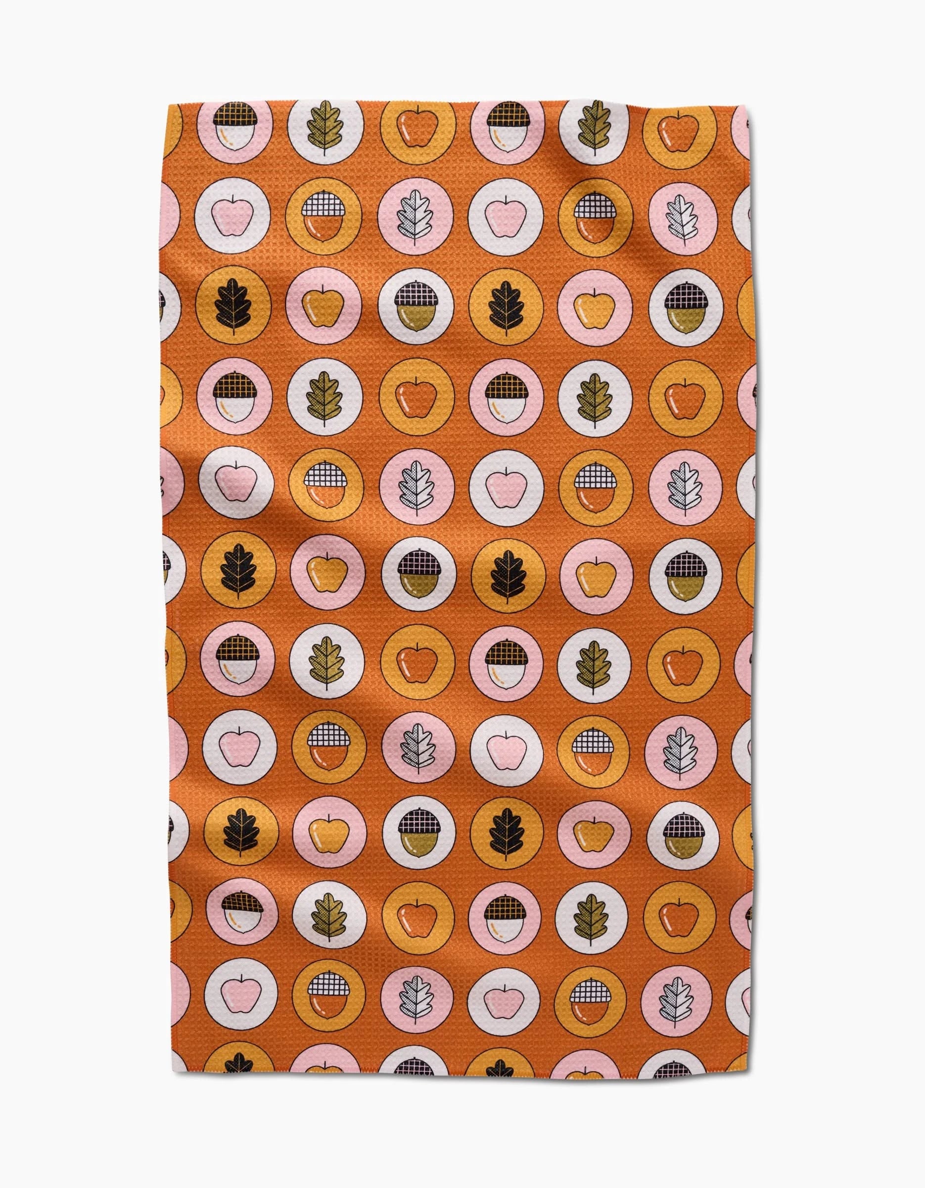 The Apples Acorns tea towel by Megan Roy is quick-drying and features a vibrant orange background with circles, apples, leaves, grid designs, and acorns in orange, pink, brown, and gold.