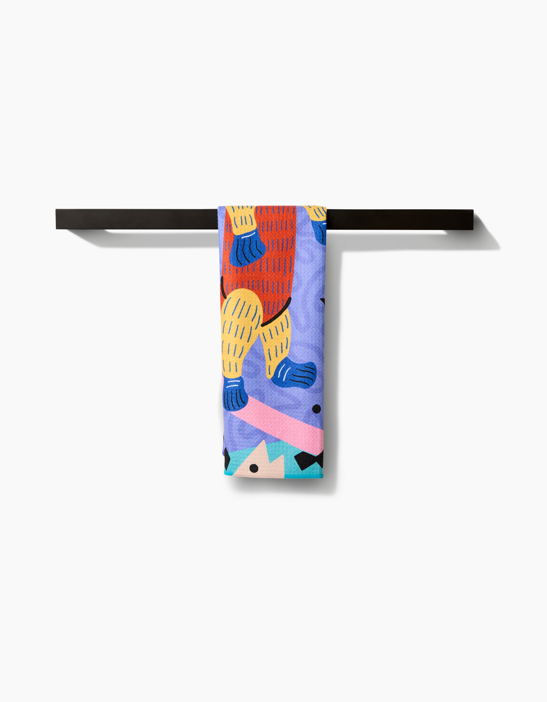 The Ariranha tea towel by Camila Leão, featuring a colorful abstract design with bold shapes in blue, red, yellow, and pink, hangs neatly on a simple black towel bar against a white background.