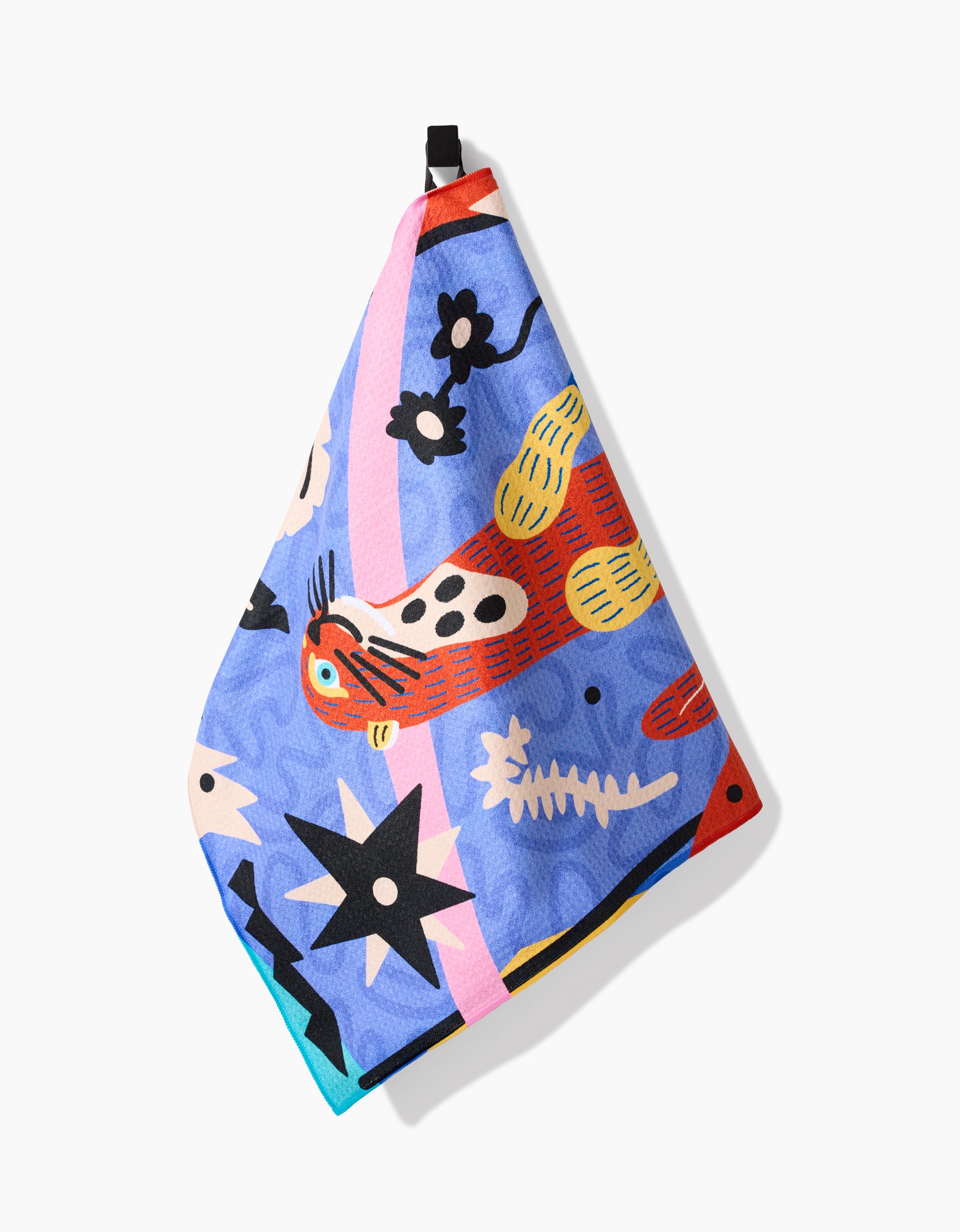 A vibrant Camila Leão Ariranha tea towel hangs on a hook, displaying bold abstract designs—spotted animal, flowers, stars, wavy lines, and a fish skeleton—on blue Geoweave fabric.