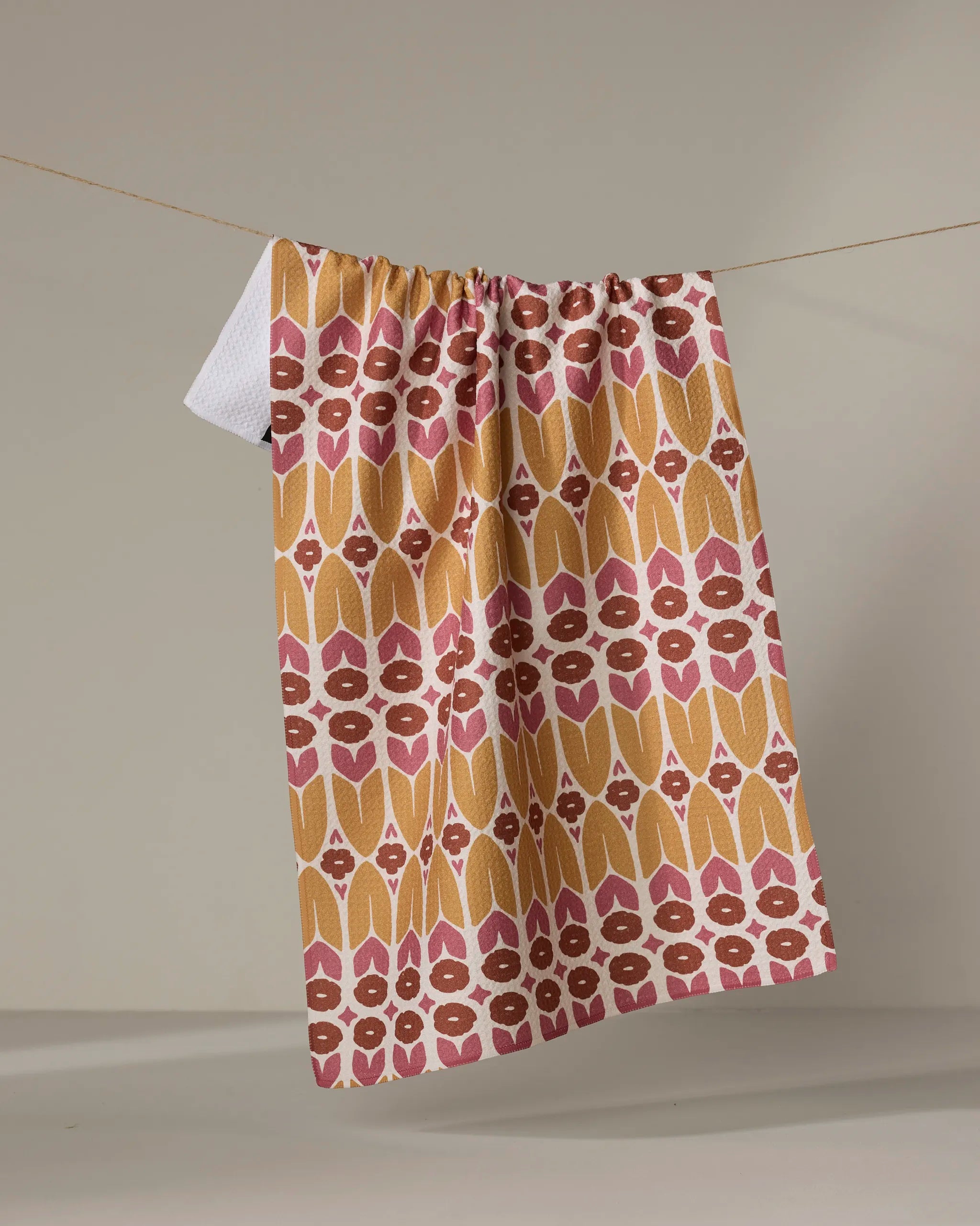The Kayla Ann "Autumn Groove" tea towel, featuring pink, mustard yellow, and white geometric shapes, hangs on a clothesline against a simple, neutral background.