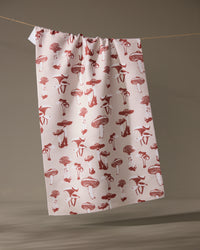 The Geometry Autumn Mushrooms tea towel, made from recycled materials and featuring a red mushroom and animal pattern, hangs on a string against a neutral background.