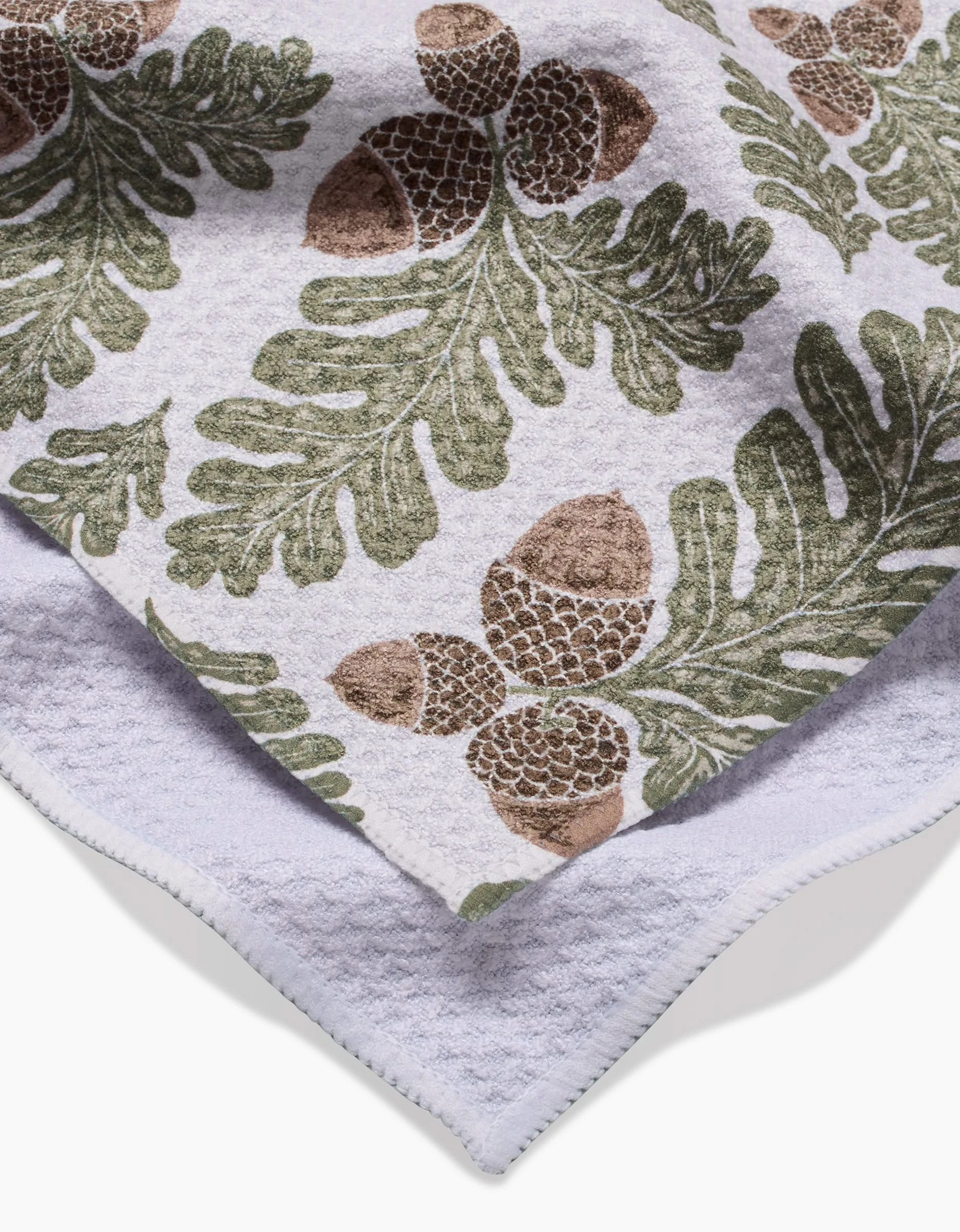 The Autumn Pines towel by Danelys Sidron features ultra-absorbent Geoweave fabric with a green oak leaf and brown acorn pattern. The image shows it folded, revealing part of the plain underside.