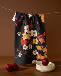 The Geometry Autumn Whispers recycled kitchen towel, featuring a dark floral pattern with red and white flowers, hangs on a string above two white bowls and two red apples set against a brown background.