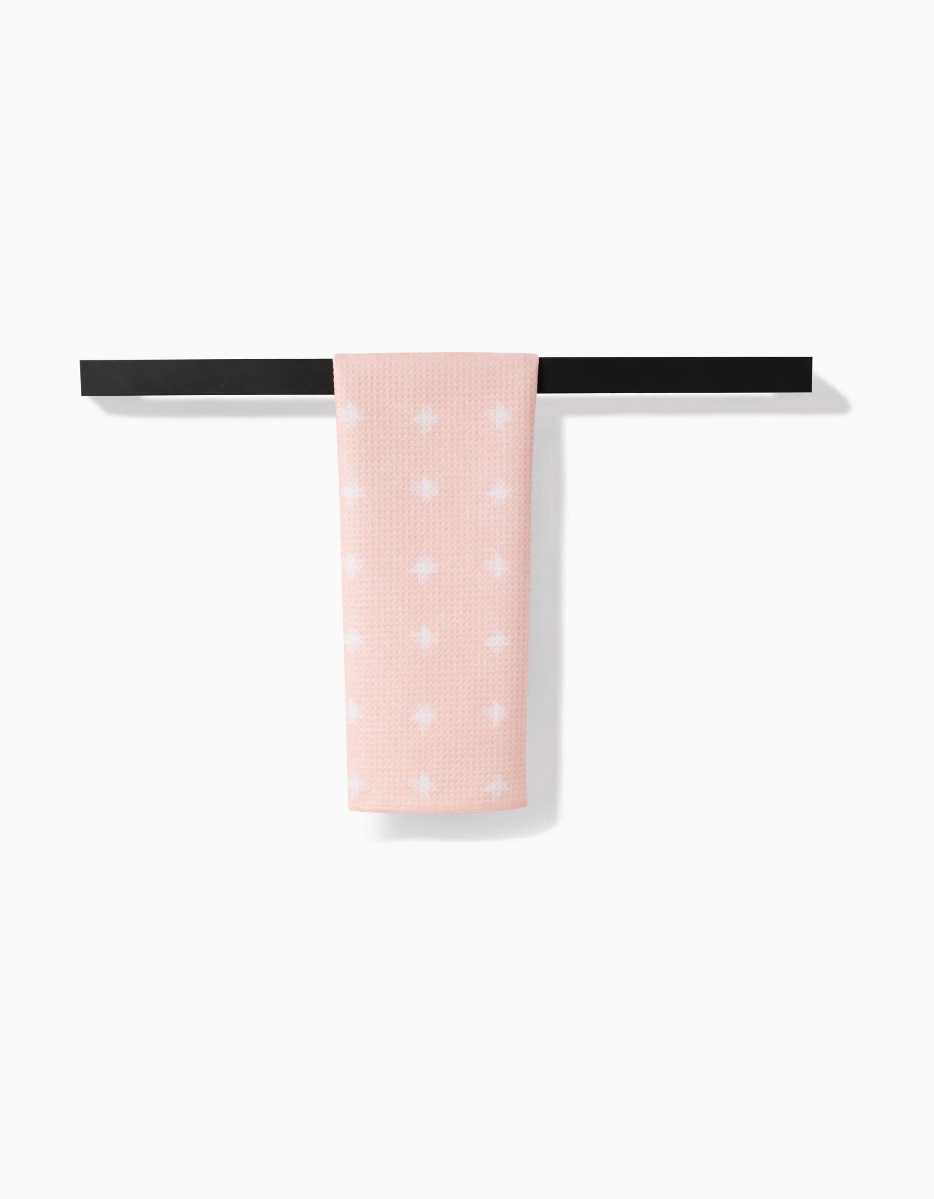 The Amy MacCready Aveline tea towel, in light pink with white polka dots and made from recycled materials, is neatly folded and draped over a sleek black towel bar against a plain white background.