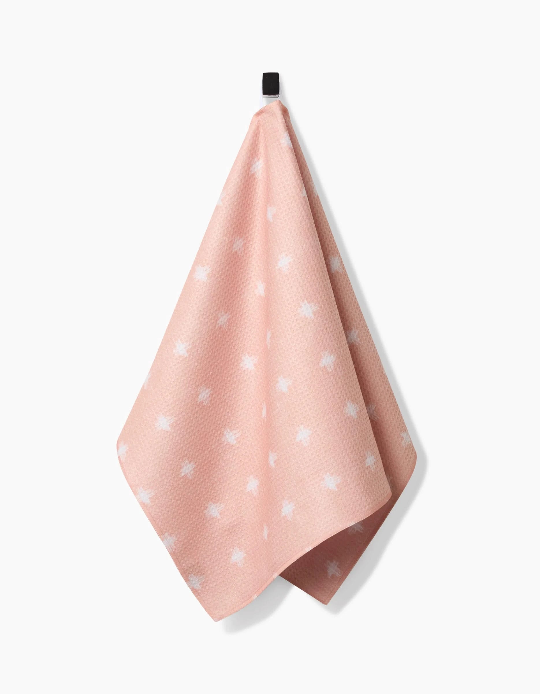 The Aveline tea towel by Amy MacCready, crafted from recycled materials, features a pink textured surface with white star-like patterns and hangs from a black hook against a white background.