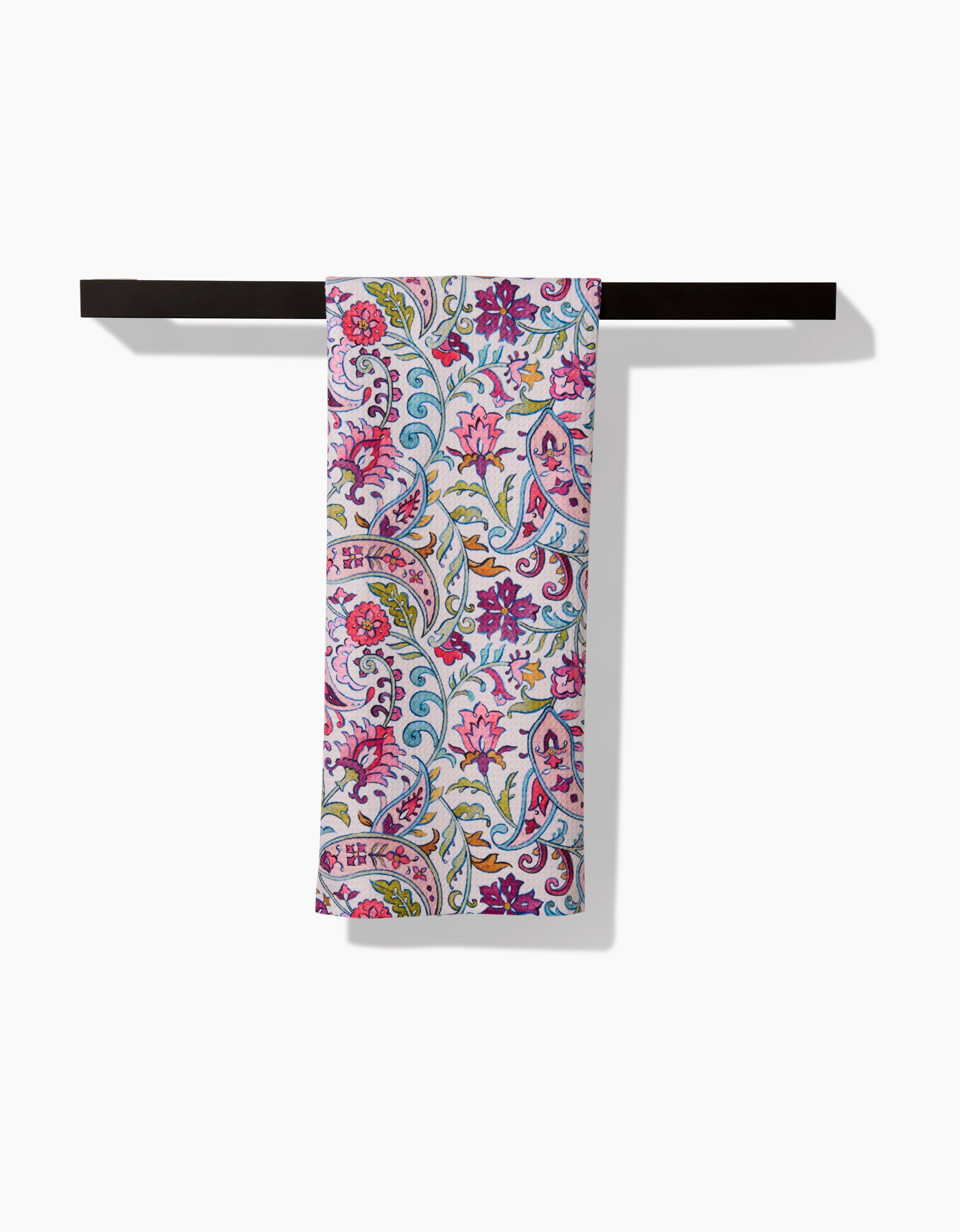 The Bazaar kitchen tea towel by Maayan Alper-Swan features a vibrant floral and paisley pattern in pink, purple, blue, and green on white. Made from soft Geoweave fabric for excellent absorbency, it hangs neatly on a dark towel bar against a white background.