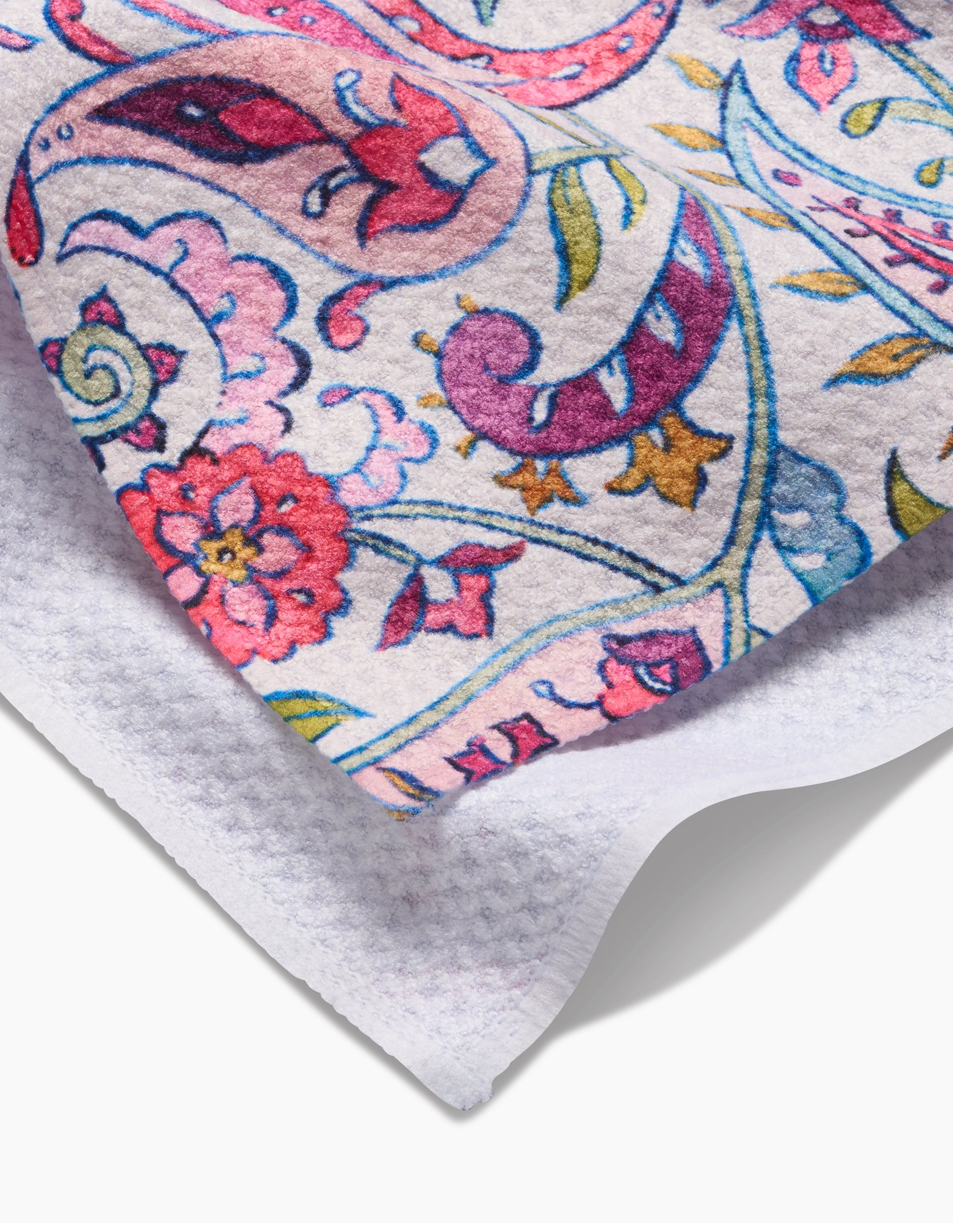 A close-up of Maayan Alper-Swans Bazaar Kitchen Tea Towel shows its textured white fabric with a vibrant paisley and floral design in pink, purple, blue, green, and gold. One corner is folded to display the plain white reverse side.