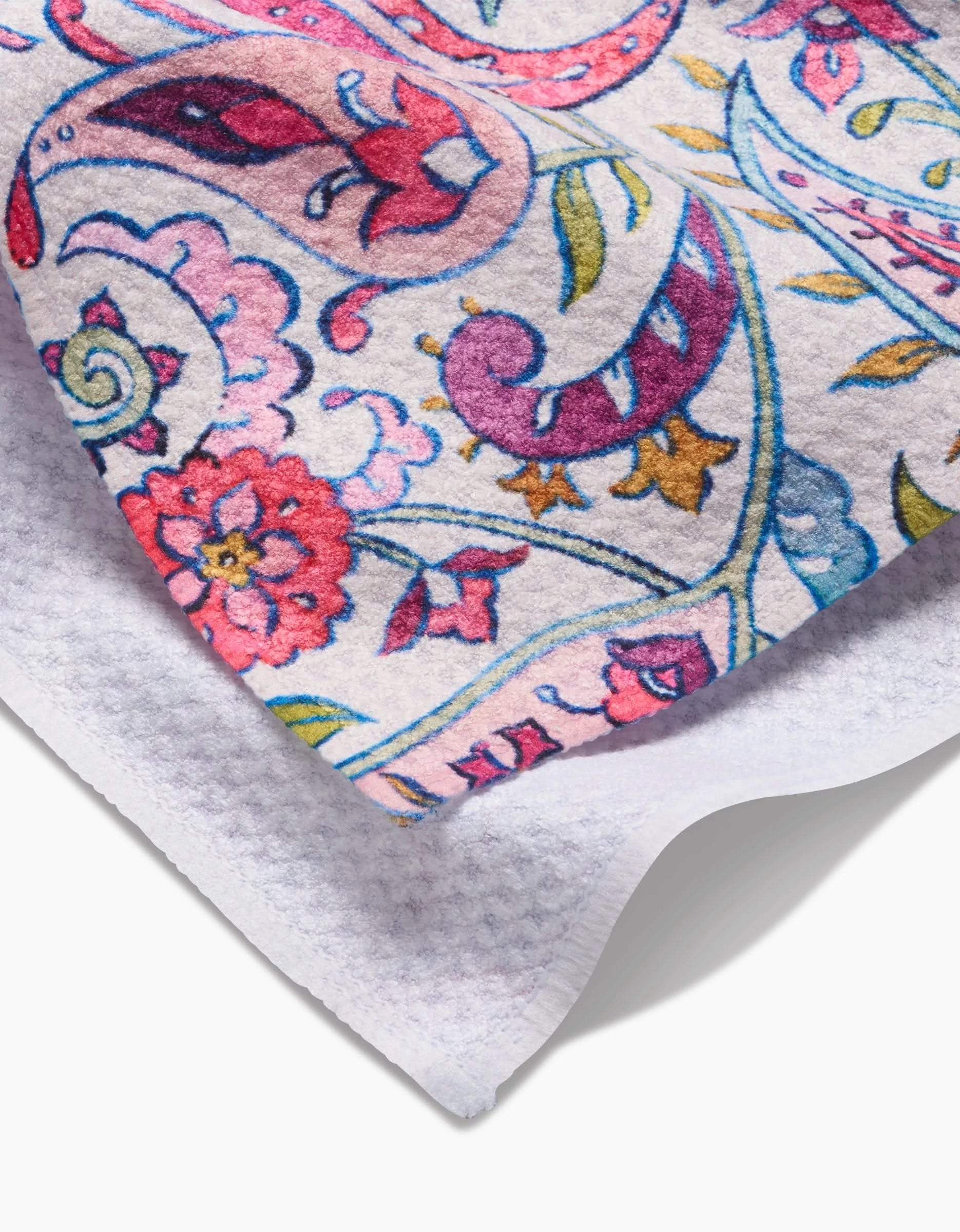 A close-up of Maayan Alper-Swans Bazaar Kitchen Tea Towel shows its textured white fabric with a vibrant paisley and floral design in pink, purple, blue, green, and gold. One corner is folded to display the plain white reverse side.