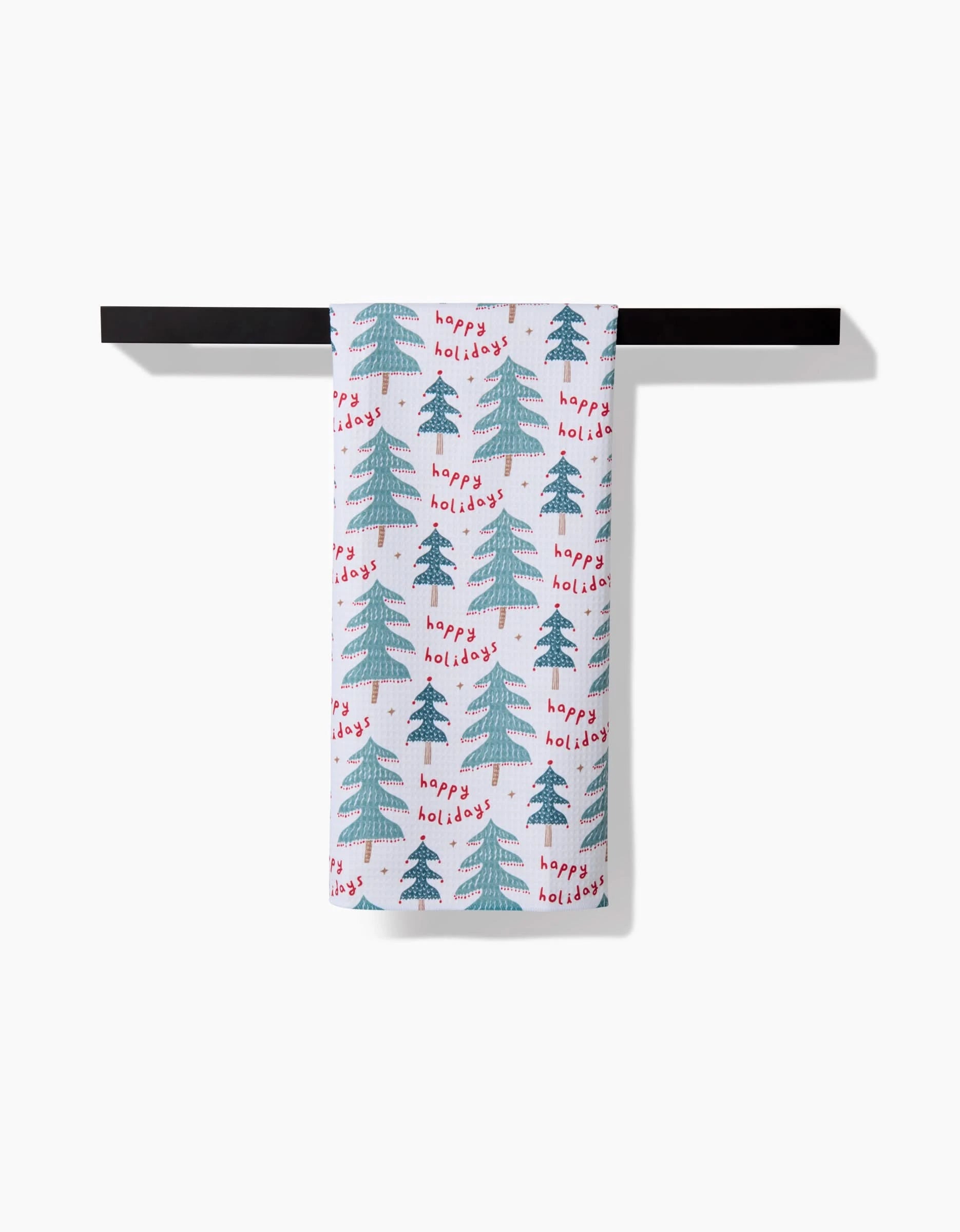 The Berry Happy Holidays kitchen towel by Roxy Marj, with green Christmas trees and red Happy Holidays text, hangs on a black horizontal bar against a white background.