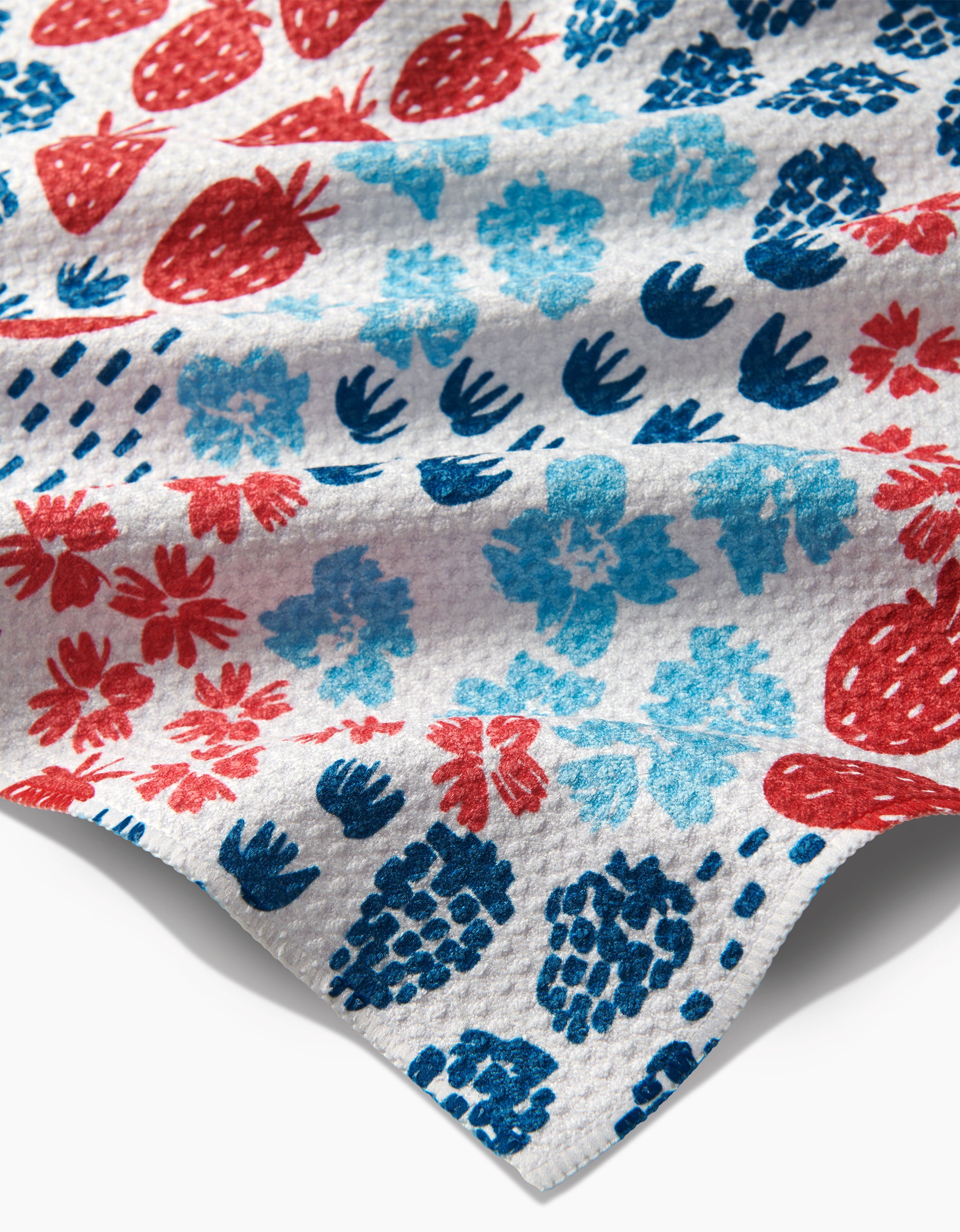 The Berry Meadow tea towel by Hufton Studio features a textured white geoweave fabric with a vibrant fruit and floral print of red strawberries, blue pineapples, and blue and red flowers.