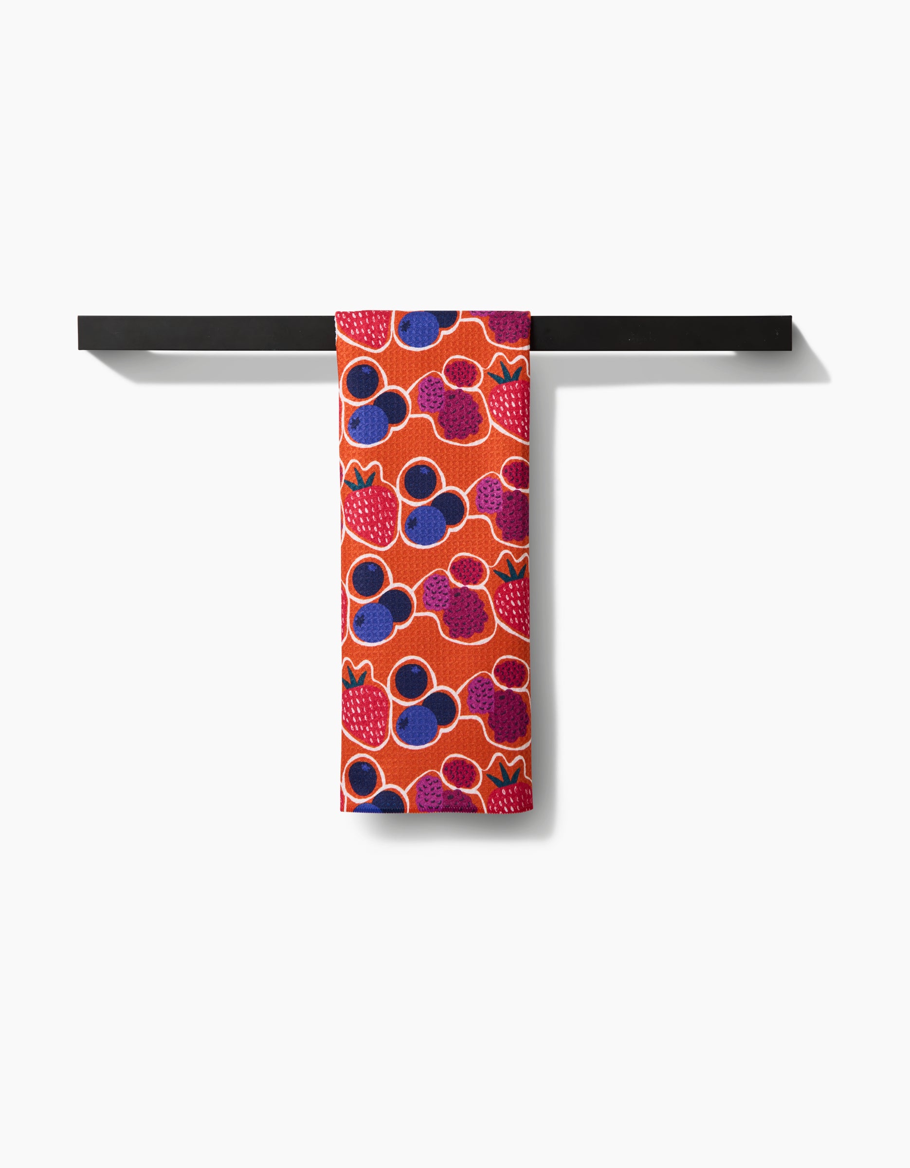 The Lizzie Clark Berrylicious tea towel, featuring a vibrant red background with strawberries and blueberries, is neatly folded over a simple black bar against a white backdrop.