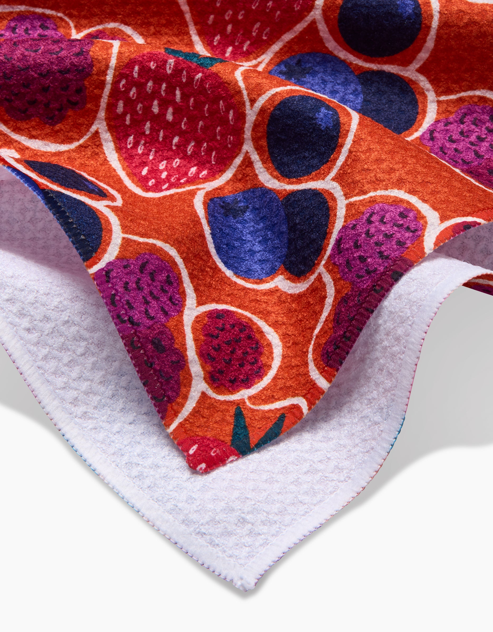 A close-up of the Lizzie Clark Berrylicious tea towel, crafted from textured Geoweave fabric and featuring a vibrant fruit pattern with strawberries, blueberries, abstract berries on an orange background, with stitched edges and a white underside.