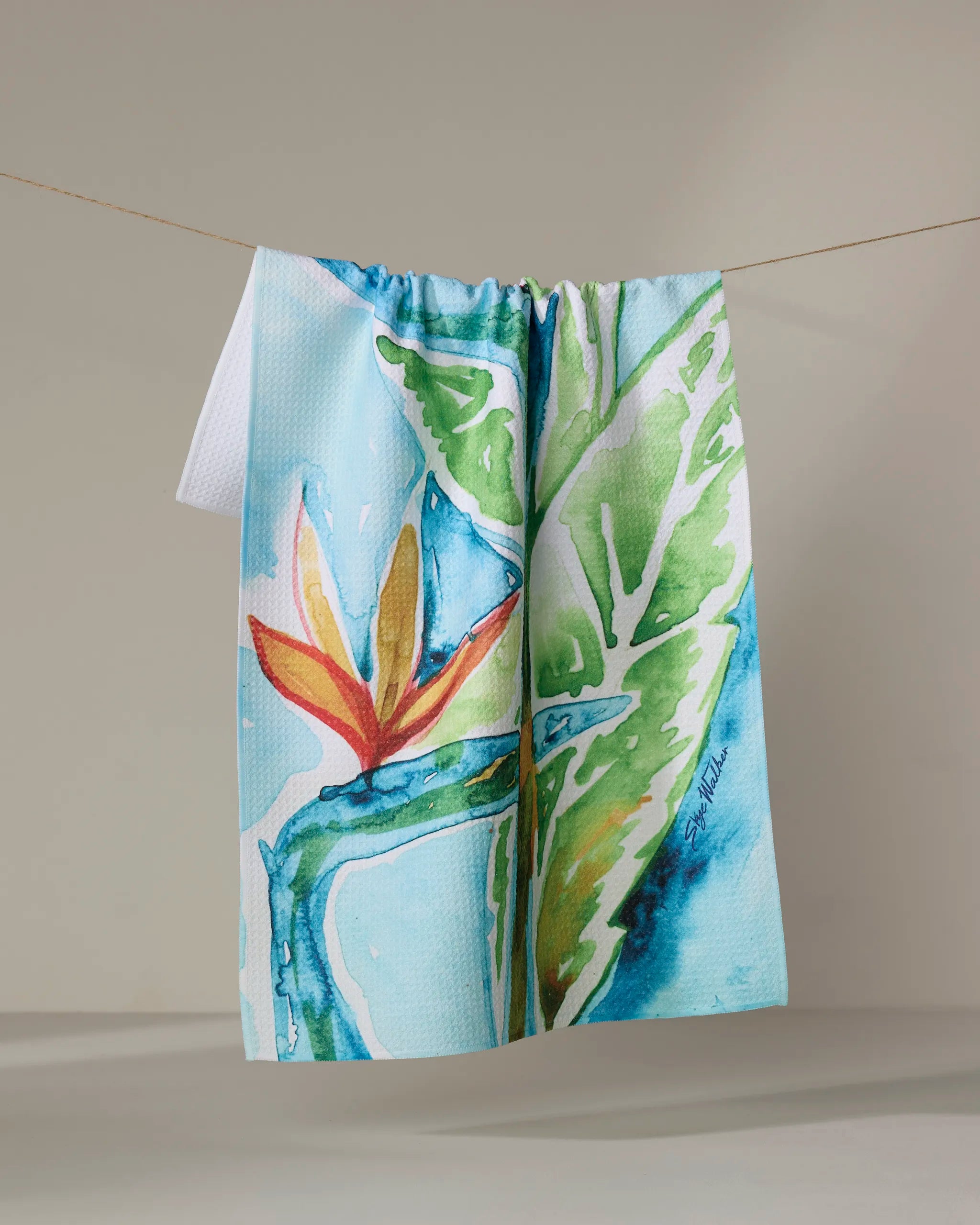 The Skye Walker "Bird Of Paradise" kitchen tea towel, crafted from recycled materials, features a vibrant watercolor green leaf and orange-red tropical flower design and hangs on a neutral background.