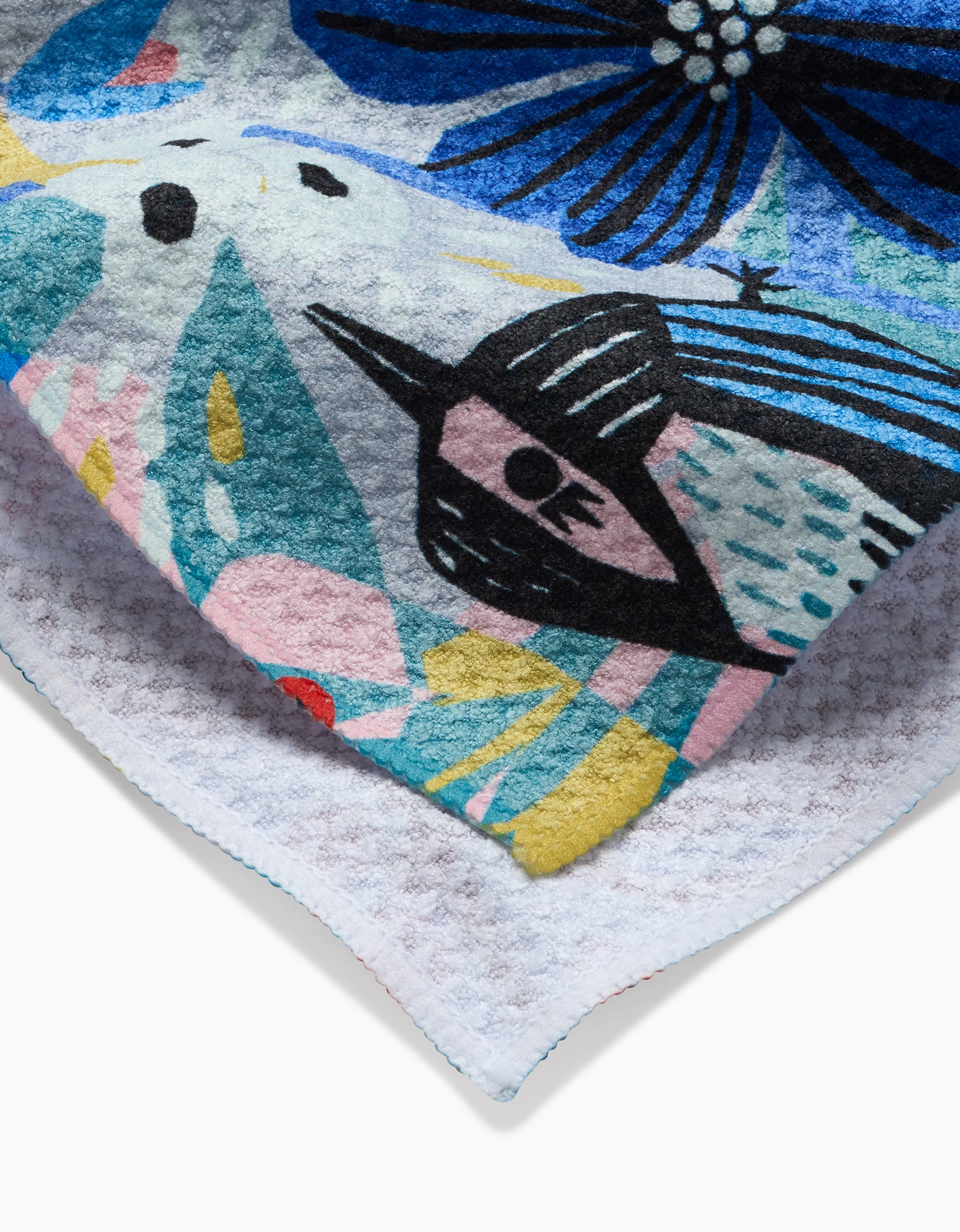 A close-up of the Birds And Berries tea towel by Masha Volnova shows abstract floral and geometric patterns in blue, black, yellow, pink, and teal. Made with Geoweave fabric, it has a textured white underside.