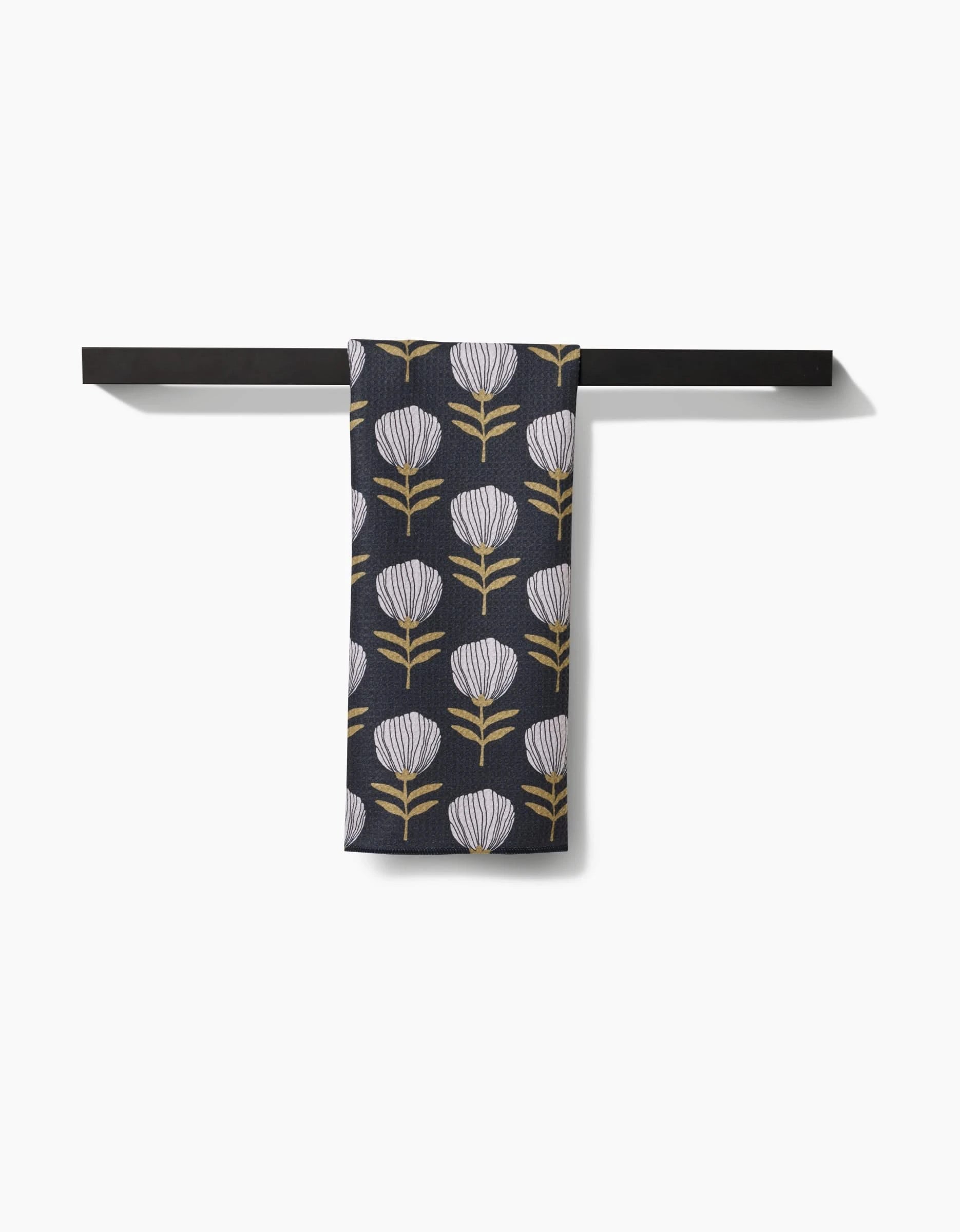 The Blomma tea towel by Anna Christian features a repeating pattern of white and gold stylized flowers on dark fabric, hangs neatly on a black towel bar, and is expertly crafted from recycled materials.