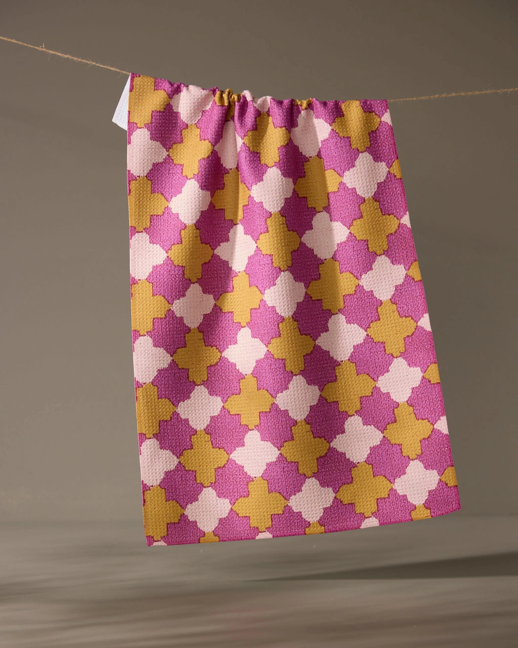 The Geometry Bloomgrid kitchen tea towel, featuring a pink, yellow, and white geometric pattern, hangs from a string against a plain neutral background.