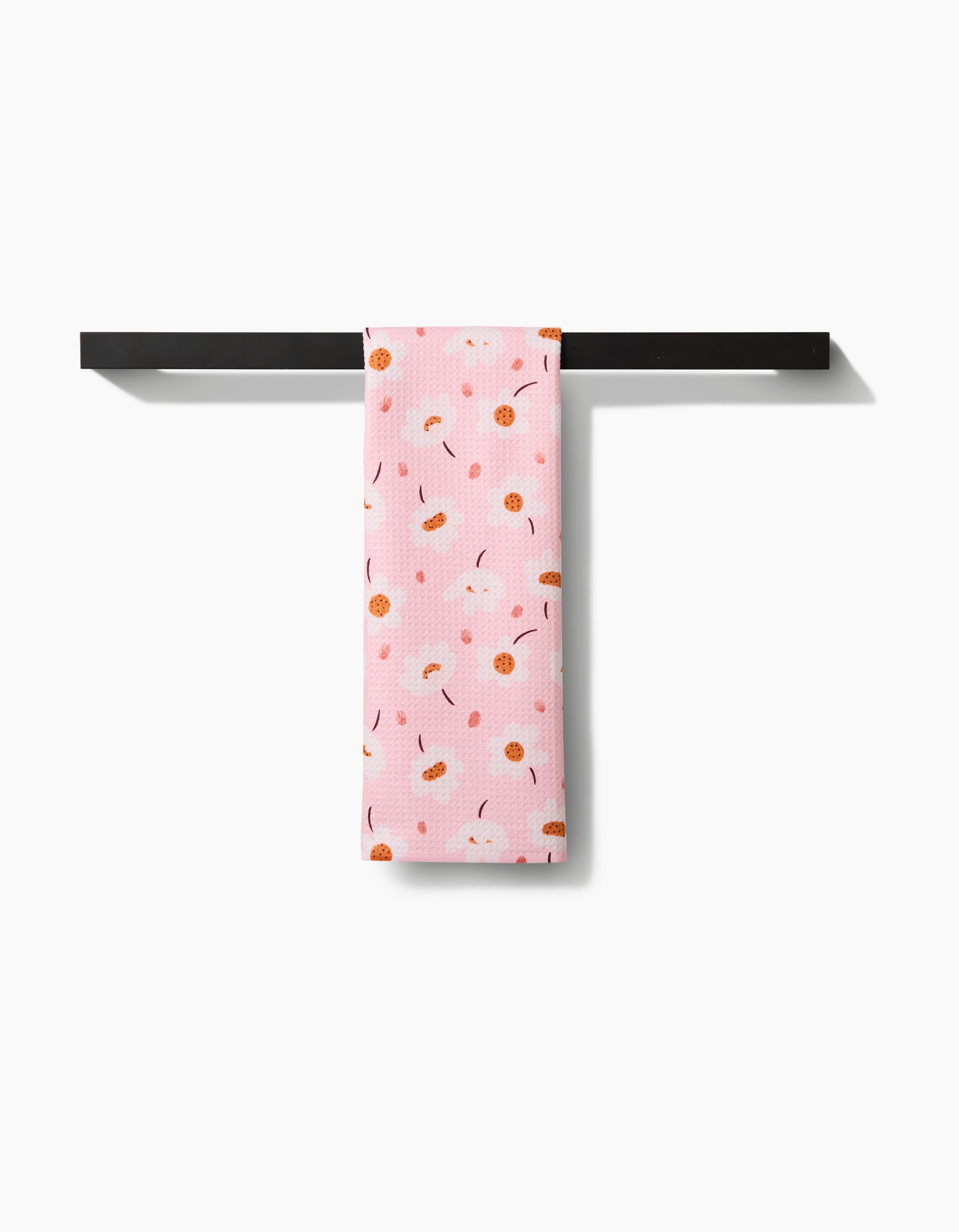 The Blush Meadows tea towel by Sara Mari, featuring an orange and white floral pattern, is neatly draped over a sleek black towel bar set against a white background.