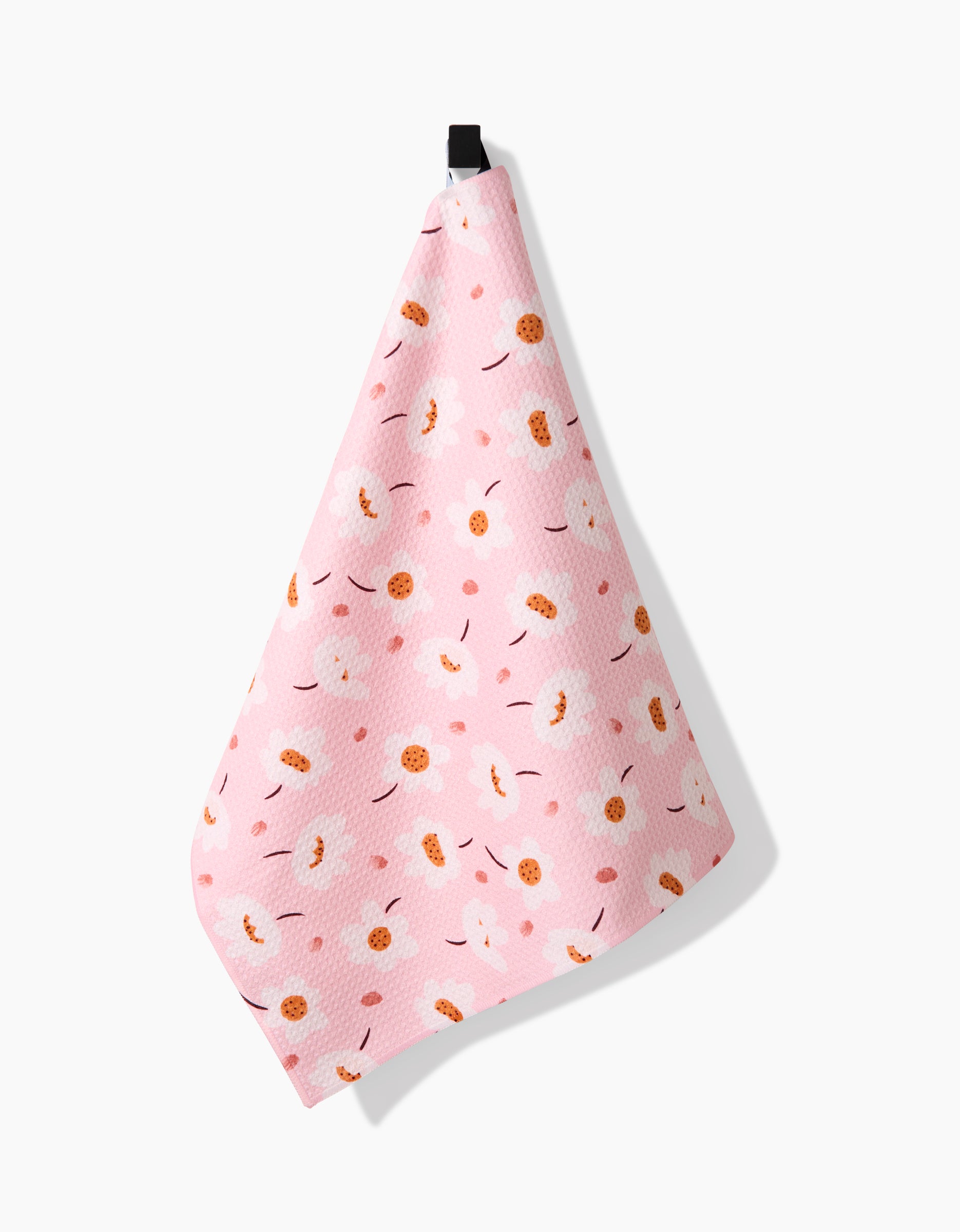 The Sara Mari Blush Meadows tea towel, crafted from Geoweave fabric, hangs from a black loop. It features a playful pattern of white clouds, orange circles, and curved brown lines on a pink background against white.