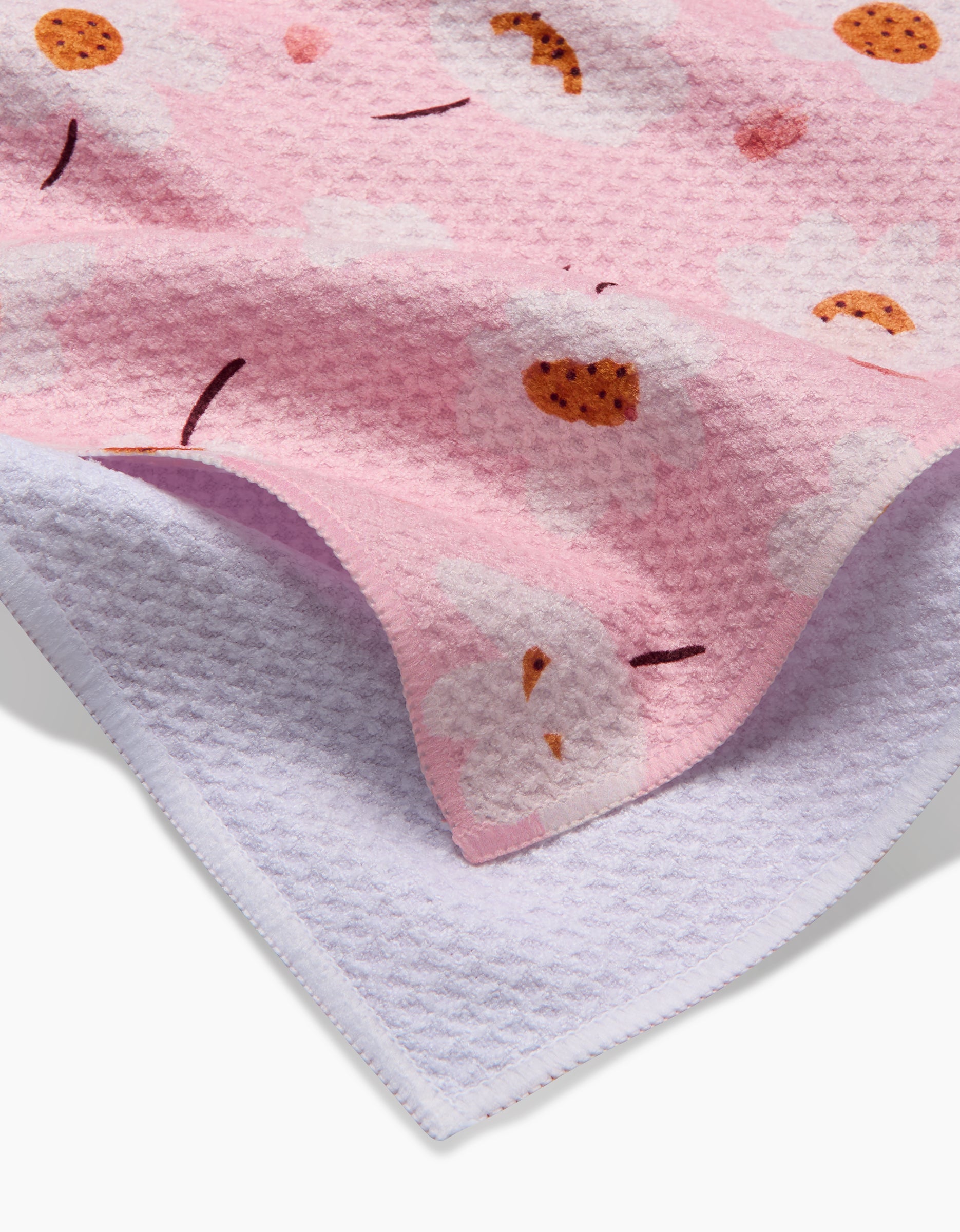 A close-up of the Sara Mari Blush Meadows tea towel, featuring pink Geoweave fabric with clouds and cookies, layered over a plain light purple fabric of similar texture.