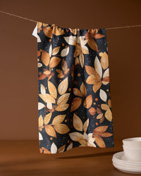 The Geometry Botanical Wonderland tea towel, made from recycled materials and featuring an autumn leaf and berry pattern in orange, yellow, and brown, hangs above stacked white dishes against a brown background.