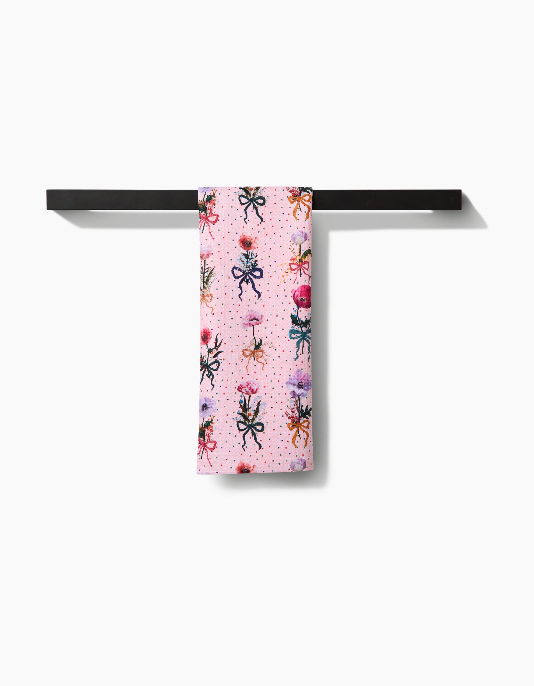 A pink Bows and Dots tea towel by Pascale Dilger, featuring a colorful insect pattern, hangs neatly over a sleek black towel bar against a white background.
