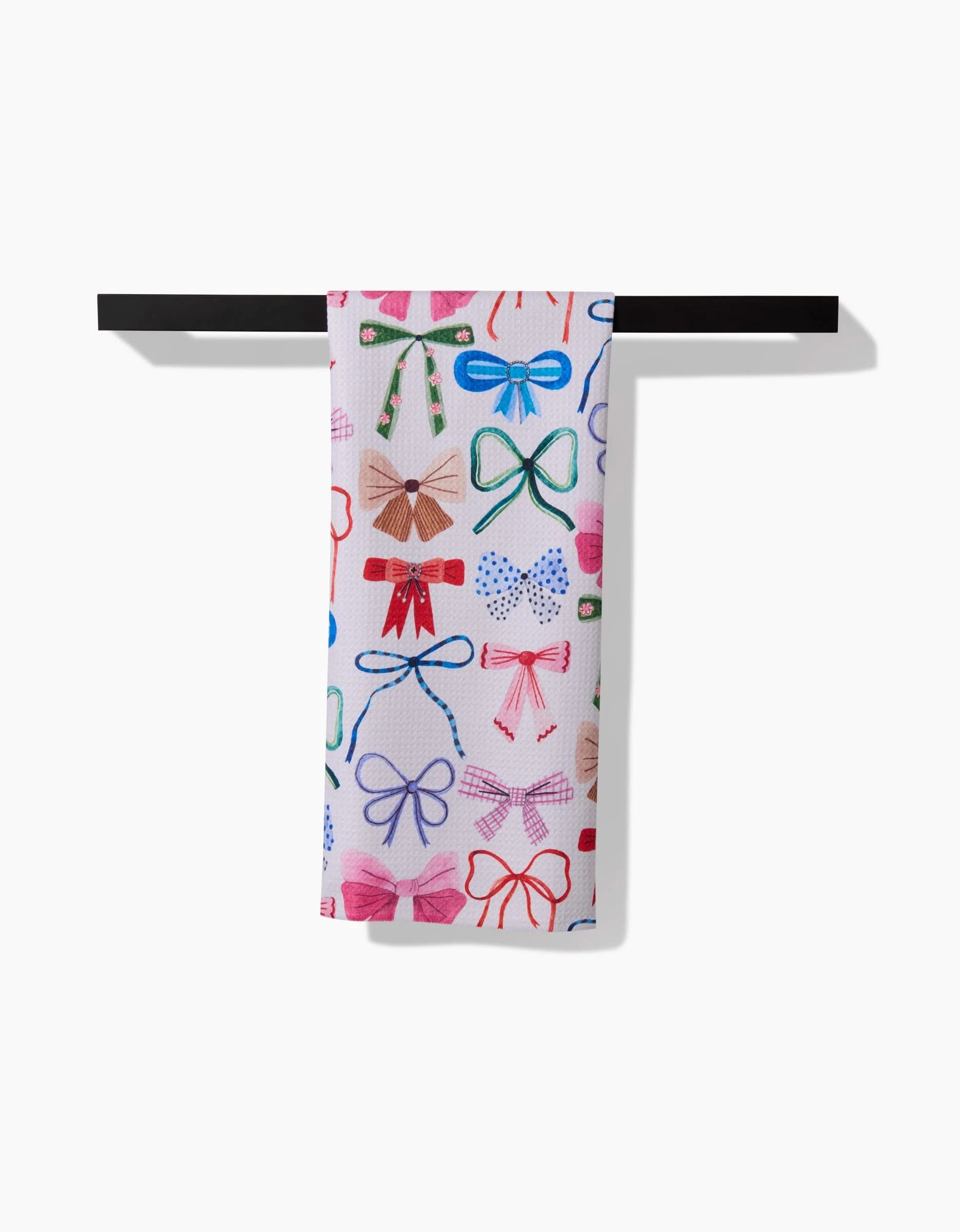 The “Bows” tea towel by Daria Solak, featuring colorful bows in various styles and sizes, hangs neatly on a black bar against a white background.