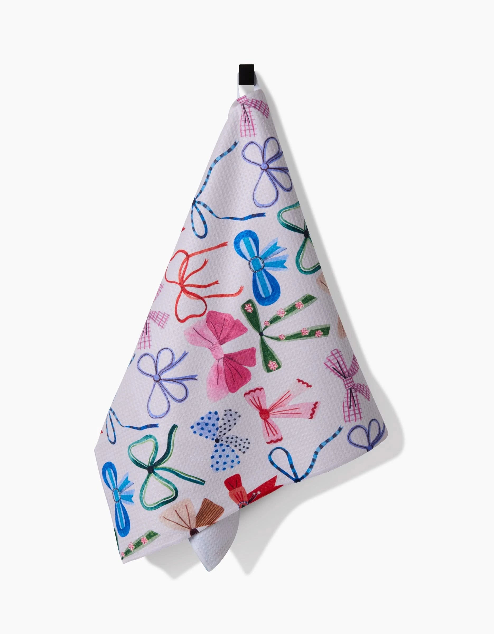 The Bows tea towel by Daria Solak features colorful red, blue, green, and pink bow patterns on white and hangs from a black hook. Made from quick-drying recycled materials, it adds stylish sustainability to your kitchen.