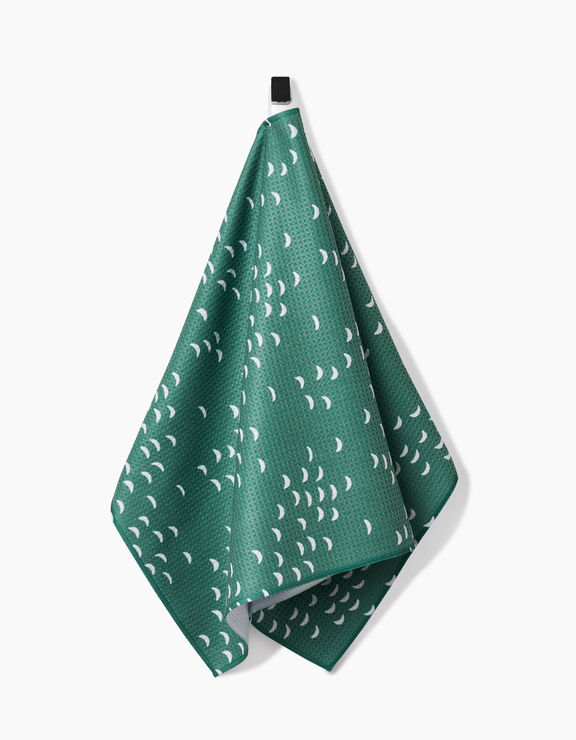 A green textured Breezeway tea towel by Jessica Swift, made from ultra-absorbent Geoweave fabric with small white crescent shapes, hangs from a black loop against a white background.