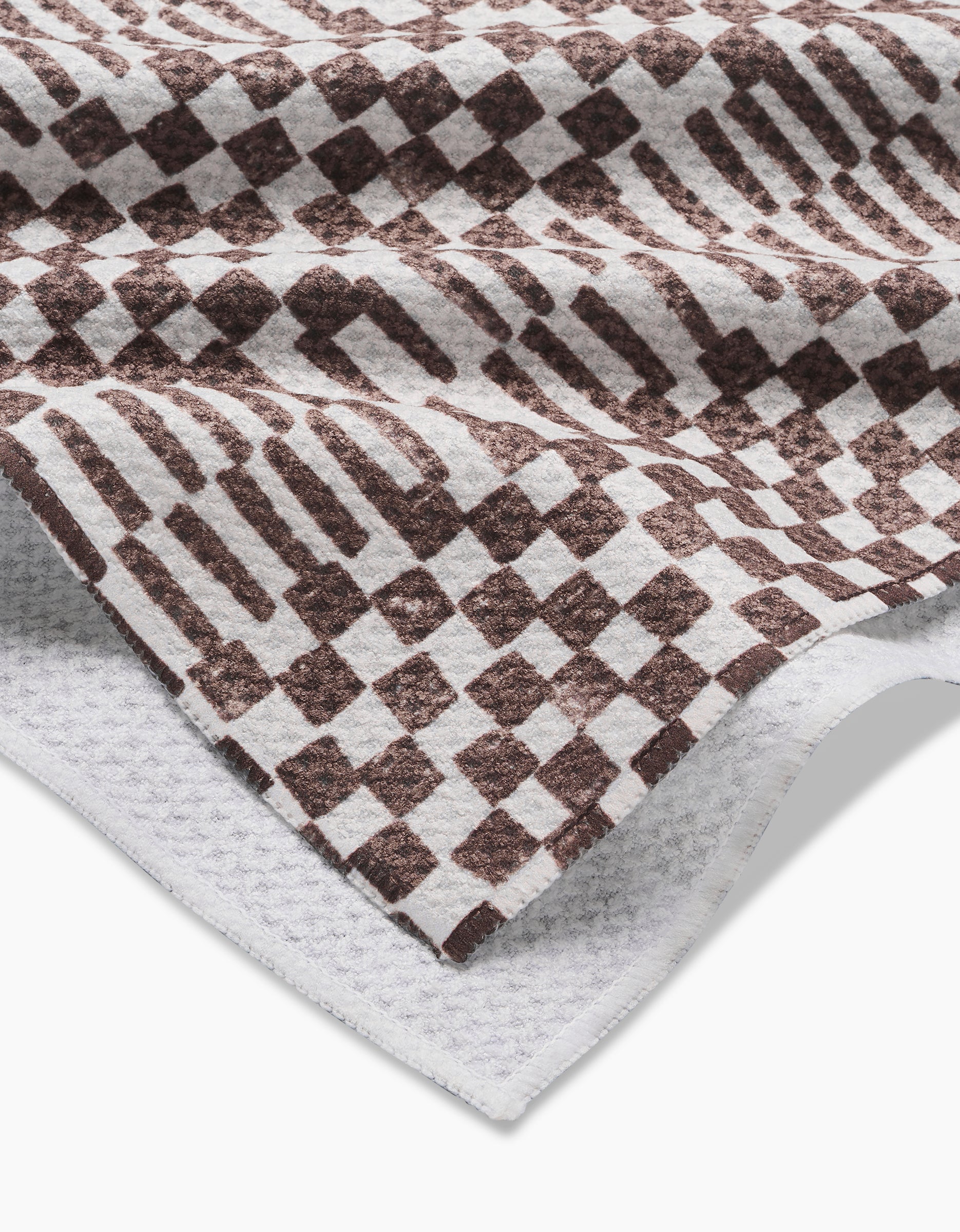 A close-up of a folded Geoweave fabric with a brown checkered and geometric pattern on white, showing its texture and edges—featured in the Studio Jeannot Chutes Coffee Tea Towel collection.