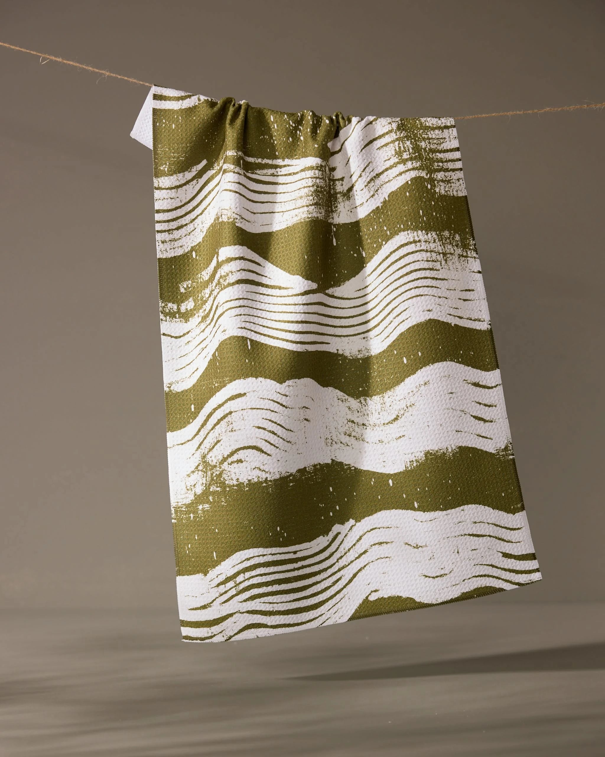The Brushline Green tea towel by Geometry, featuring green and white wavy horizontal lines, hangs from a string against a neutral background.