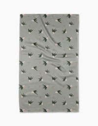 The Cali Sun Agave tea towel by Amberleigh Adoff features a grayish-green rectangular design with a subtle texture, scattered white flowers, and dark green leaves, expertly crafted from recycled materials.
