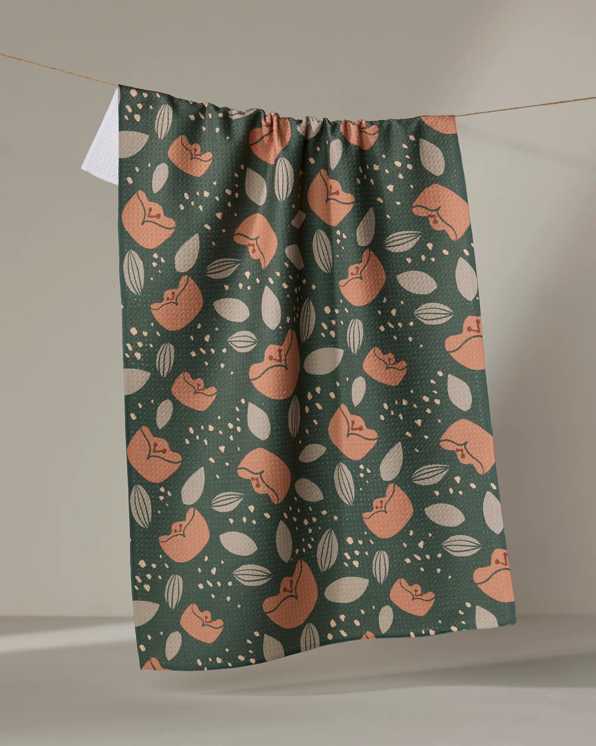 The Cali Sun tea towel by Amberleigh Adoff, made from recycled fabric, features orange foxes curled among beige leaves and dots on dark green, and hangs on a clothesline against a plain, neutral background.