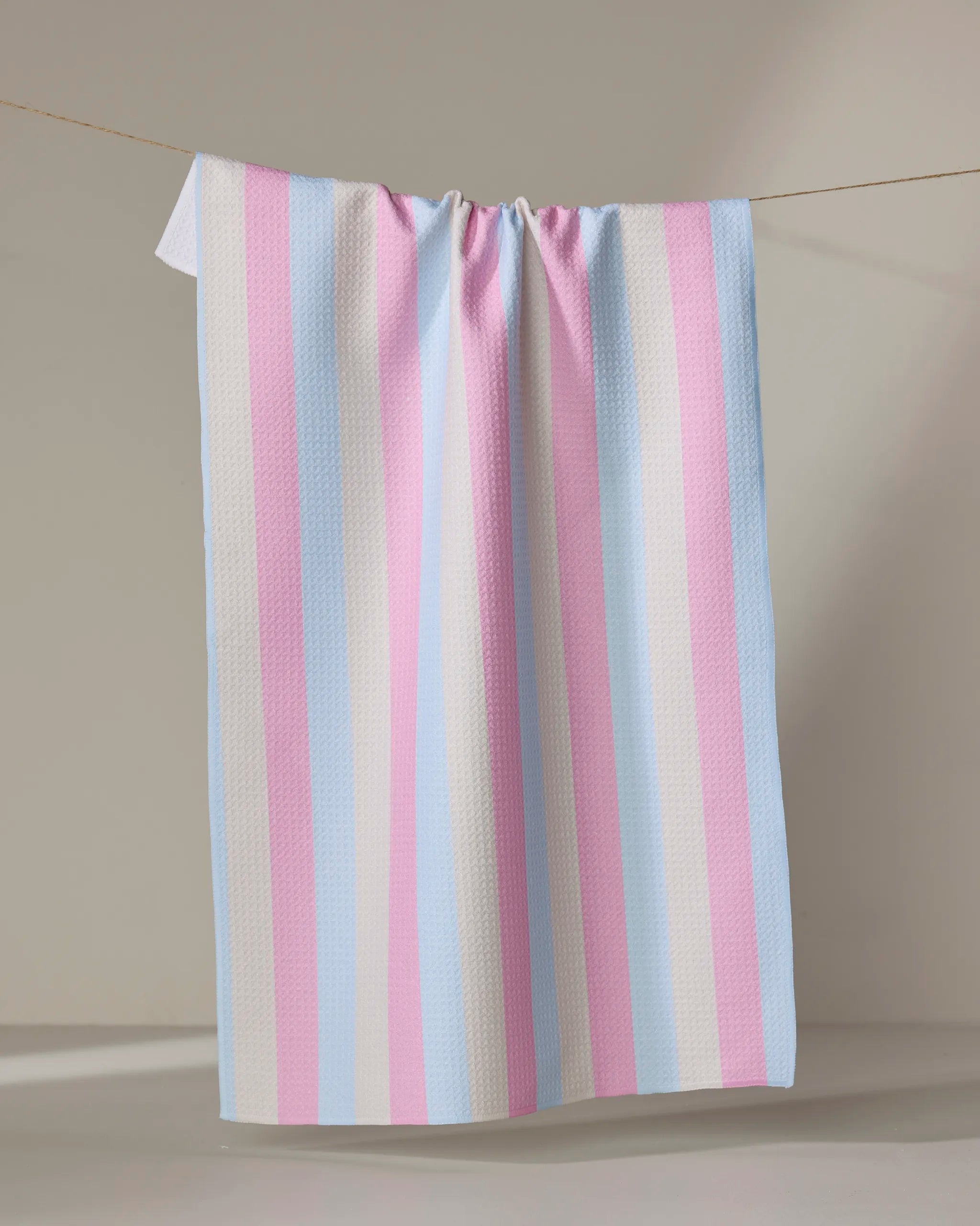 The Candy Pastels tea towel by Amy Droubay features vertical pastel stripes in light blue, pink, and white—resembling the transgender pride flag—hanging on a thin clothesline against a neutral background.