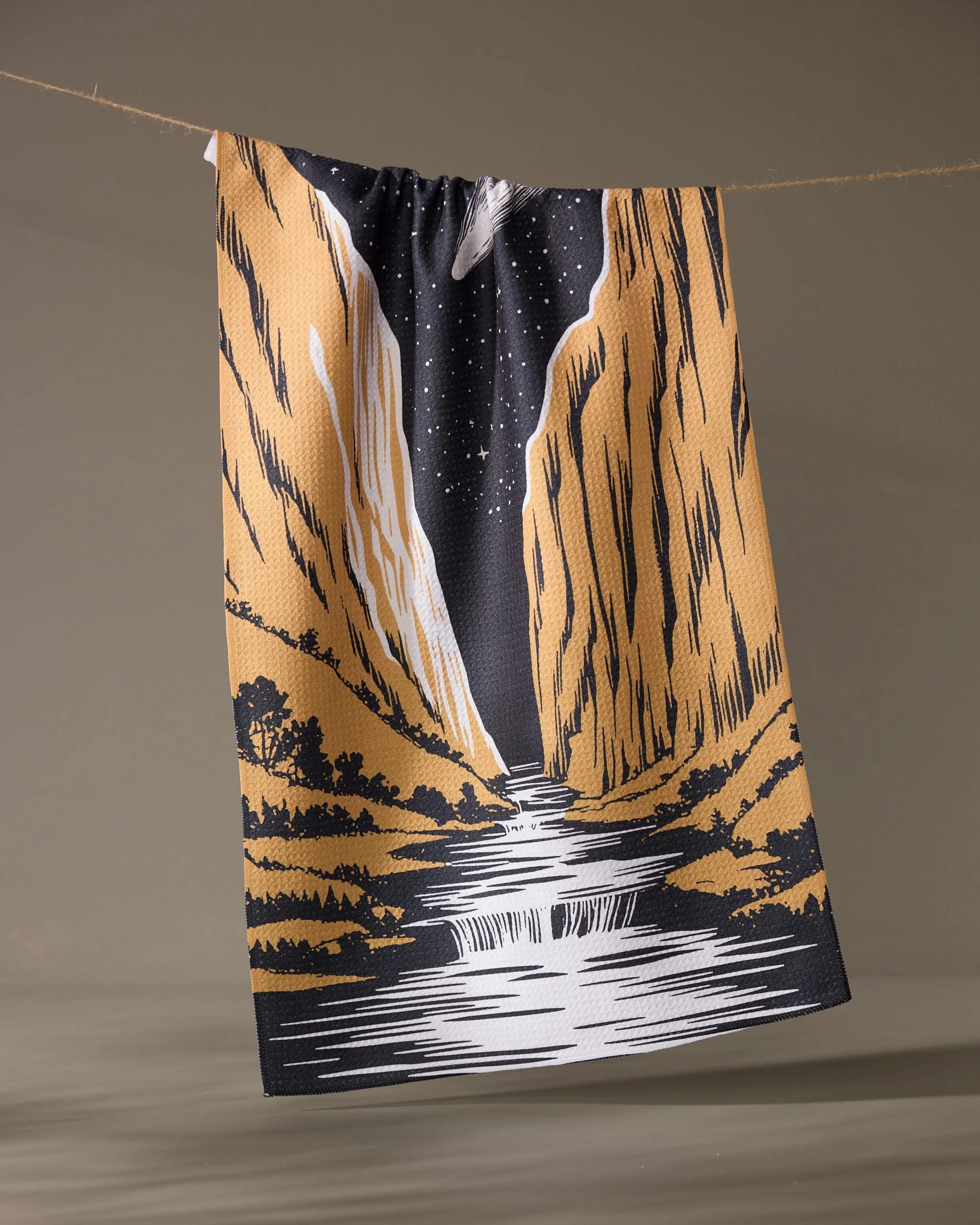 The Canyonfall kitchen towel by Geometry features an illustrated river winding between golden cliffs under a starry night sky, hanging from a clothesline against a plain background.