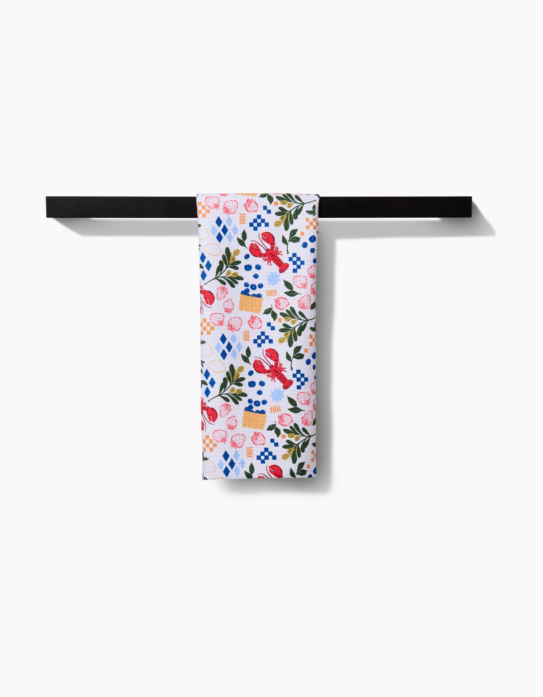 The Capri tea towel by Charlie Rowan Designs features a vibrant floral and geometric print in red, blue, pink, and green on high-quality geoweave fabric for extra absorbency. Shown draped over a minimalist black towel bar against a white background.