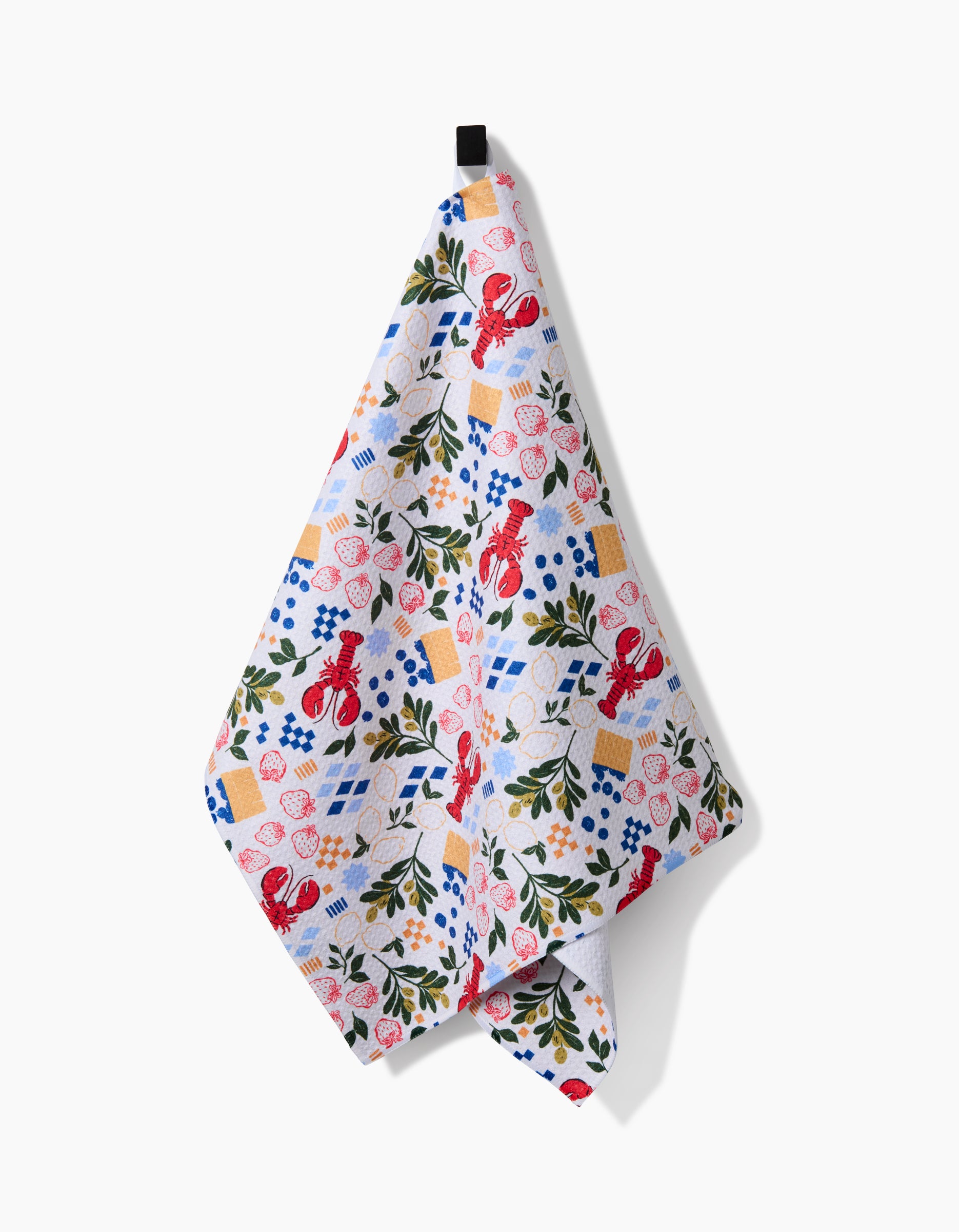 The Capri tea towel by Charlie Rowan Designs features a colorful pattern of red lobsters, lemons, blue flowers, greenery, and blue checked napkins. It hangs from a black loop and is made from recycled materials.