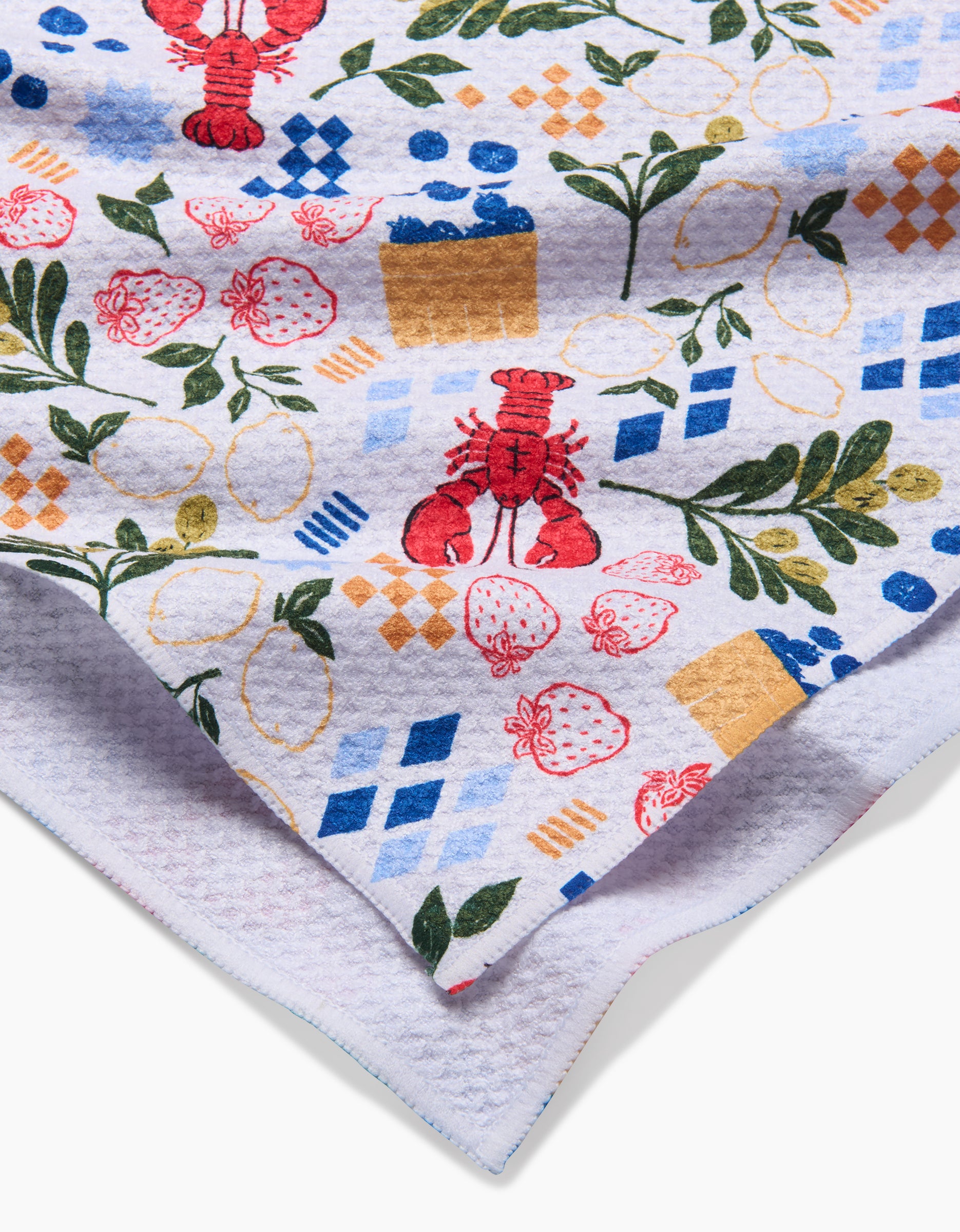 A close-up of the Capri tea towel by Charlie Rowan Designs, showcasing a white textured fabric with a colorful pattern of red lobsters, strawberries, lemons, green leaves, and blue and gold geometric shapes.