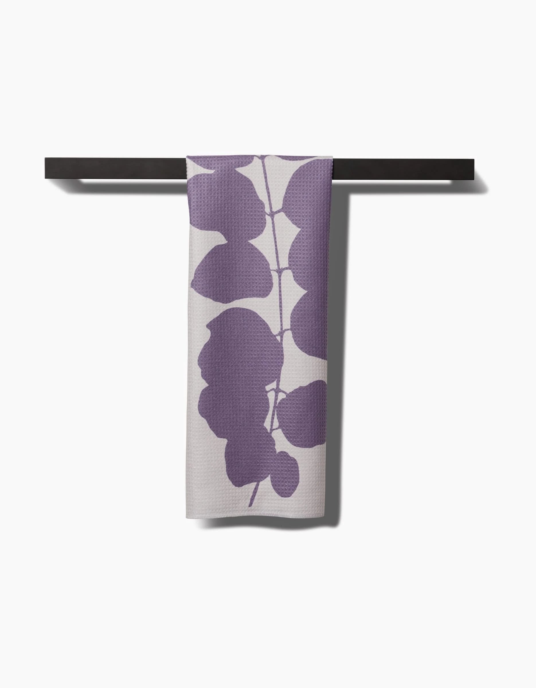 The PrintedPlant Carob towel, crafted from ultra-absorbent Geoweave fabric, features bold purple leaf patterns and hangs neatly on a dark bar against a plain white background.